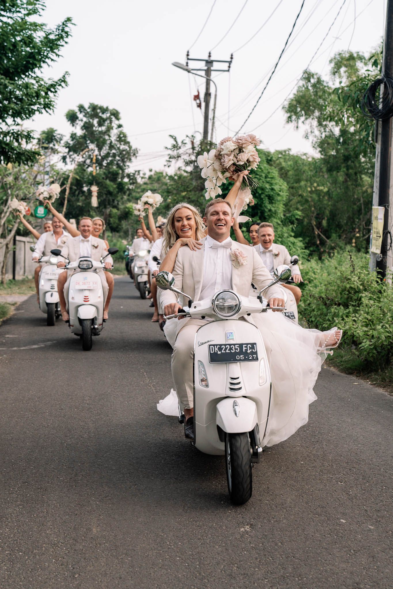 Wedding party riding white Vespa scooters down a tree-lined road in matching outfits.