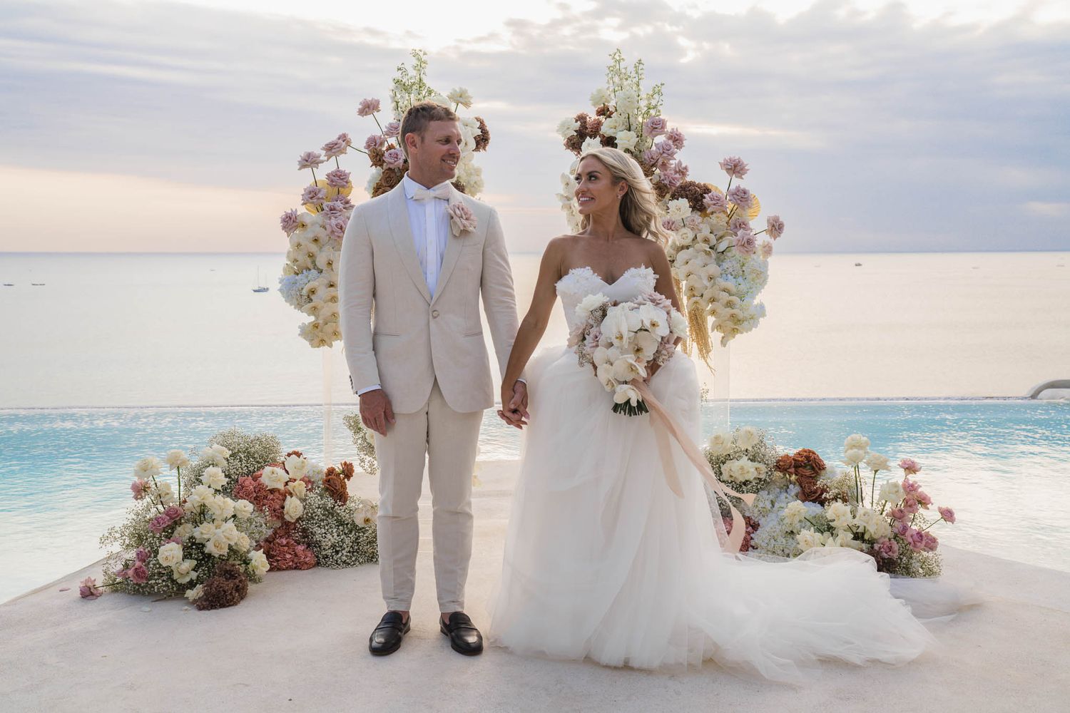 A romantic beach wedding ceremony with pink and white floral arrangements set against the turquoise ocean at sunset.