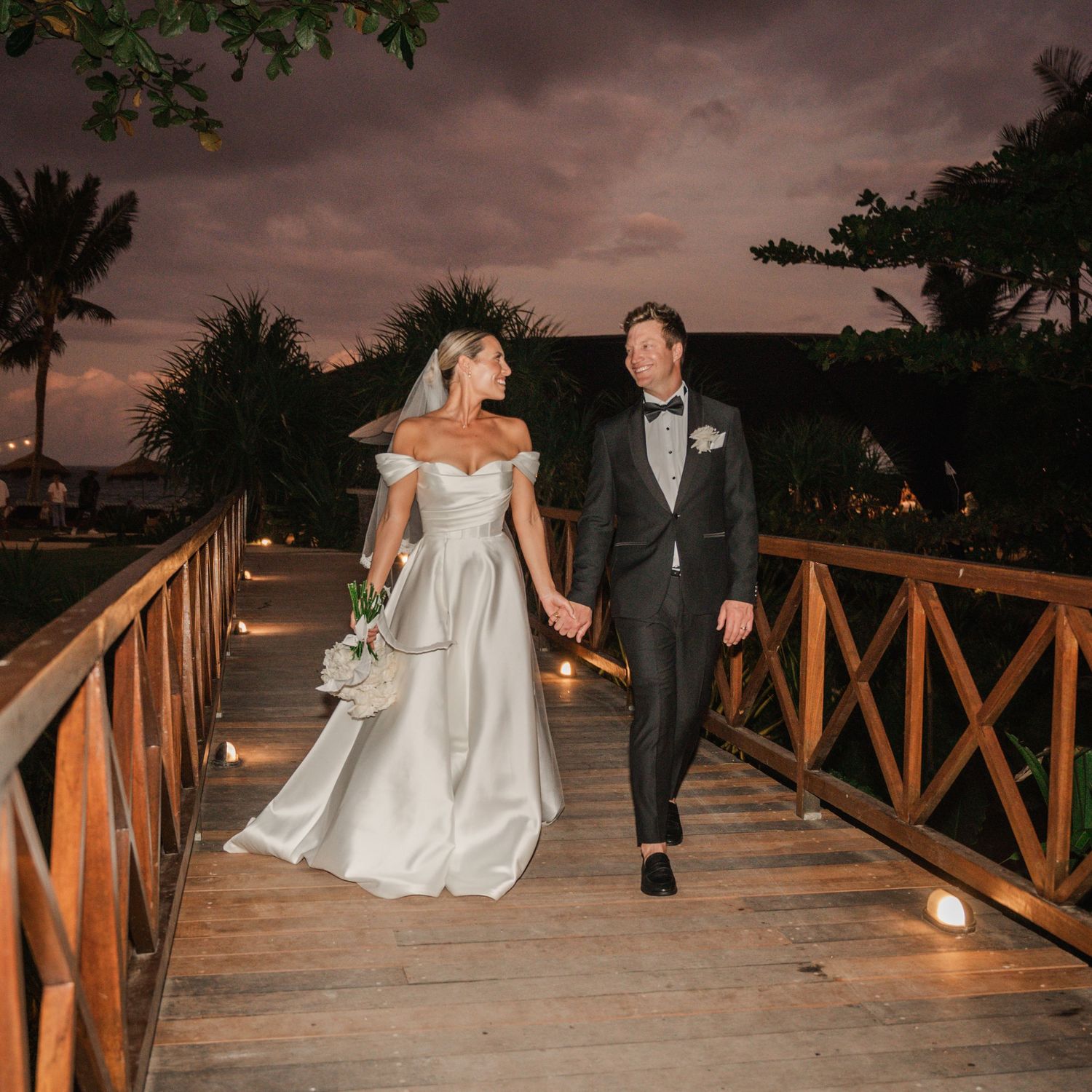 Newlyweds walk along a wooden boardwalk at sunset creating a romantic silhouette against the dramatic sky.