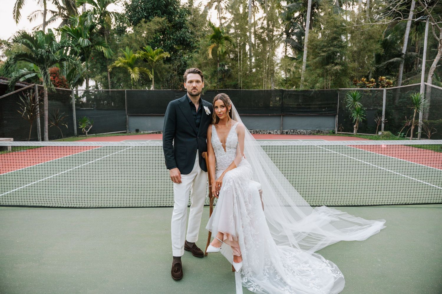 A wedding couple poses on a tennis court surrounded by palm trees and greenery.