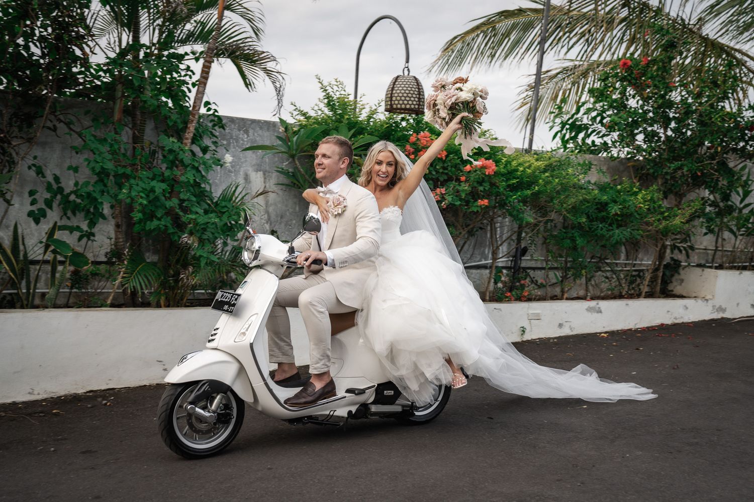 Newlywed couple poses playfully with vintage motorcycle among lush tropical garden setting.