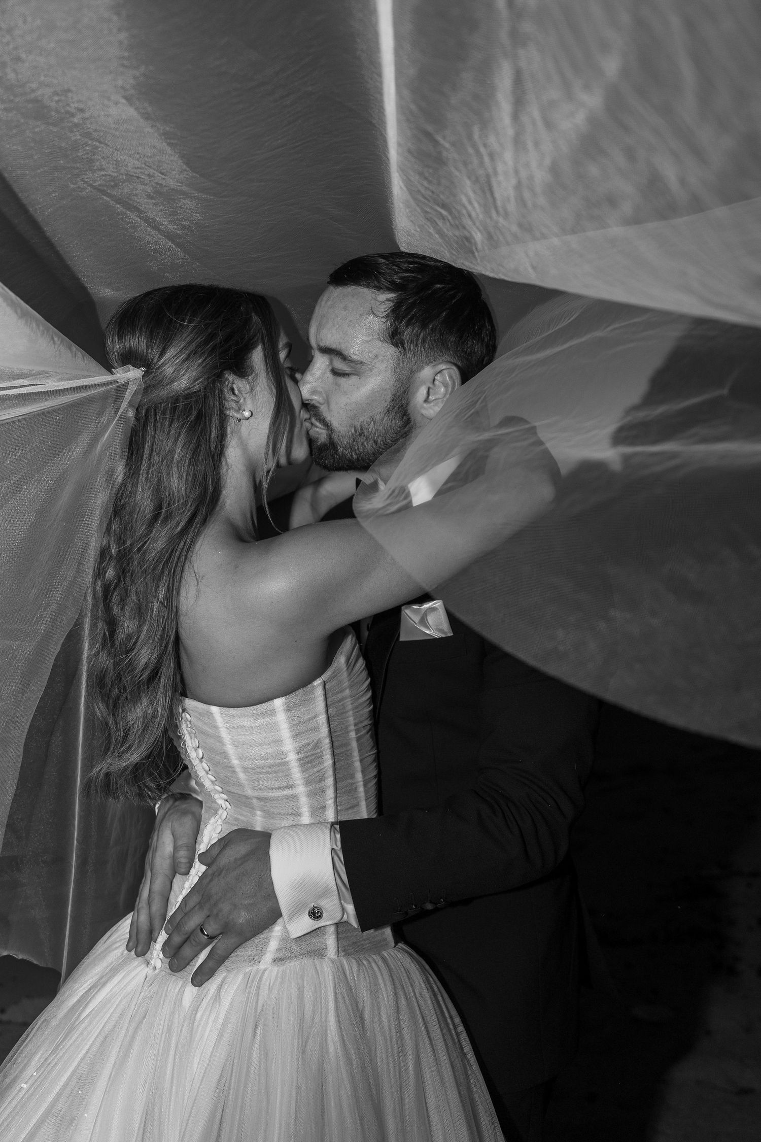 A romantic moment under a flowing white veil as newlyweds share an intimate kiss.
