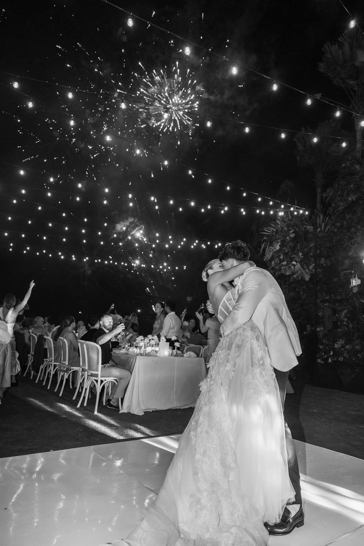 A couple shares their first dance under twinkling lights and fireworks at an outdoor evening wedding reception.