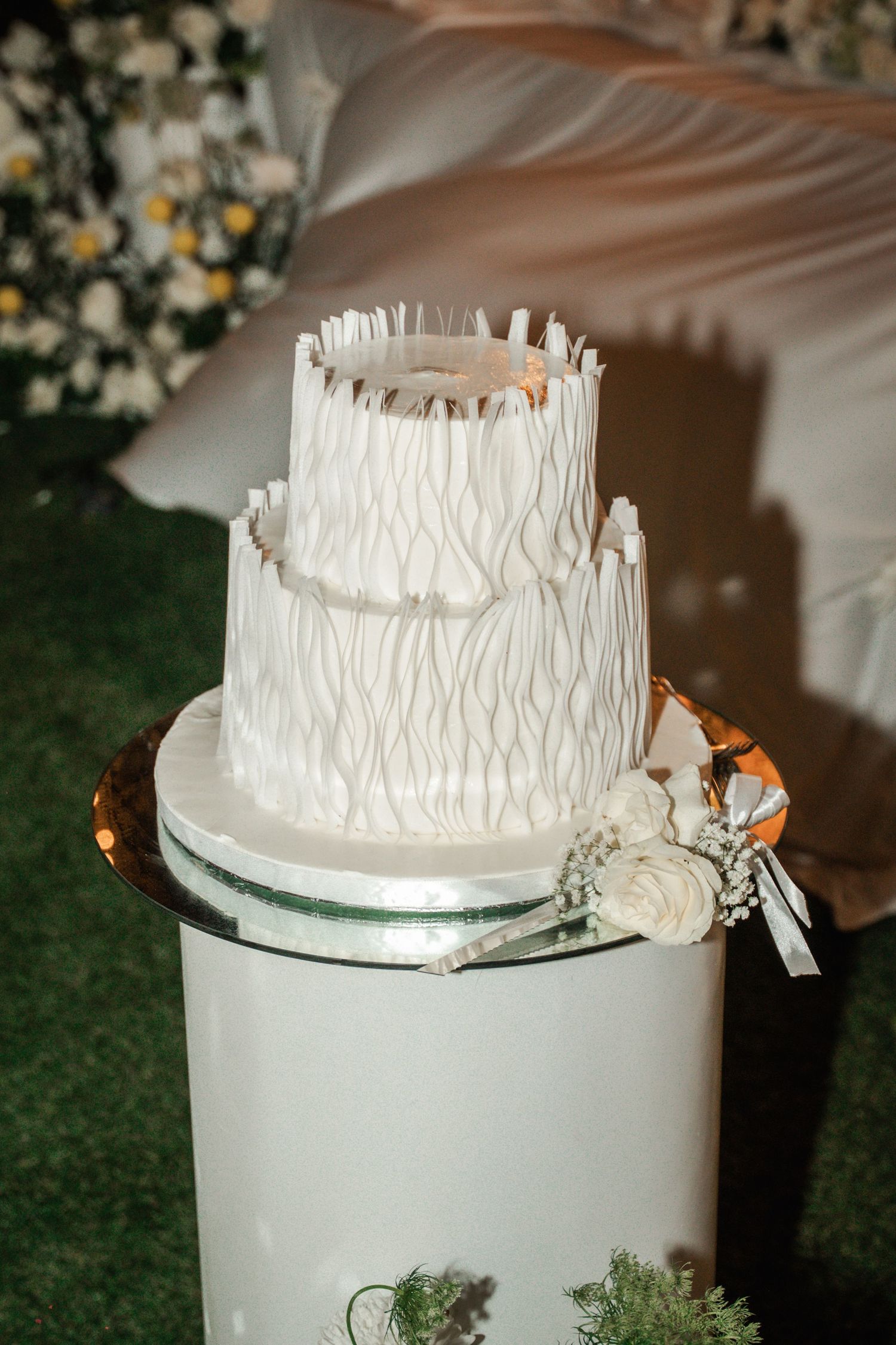 White wedding cake with textured frosting and metallic accents sits on a mirrored display stand at an outdoor reception.