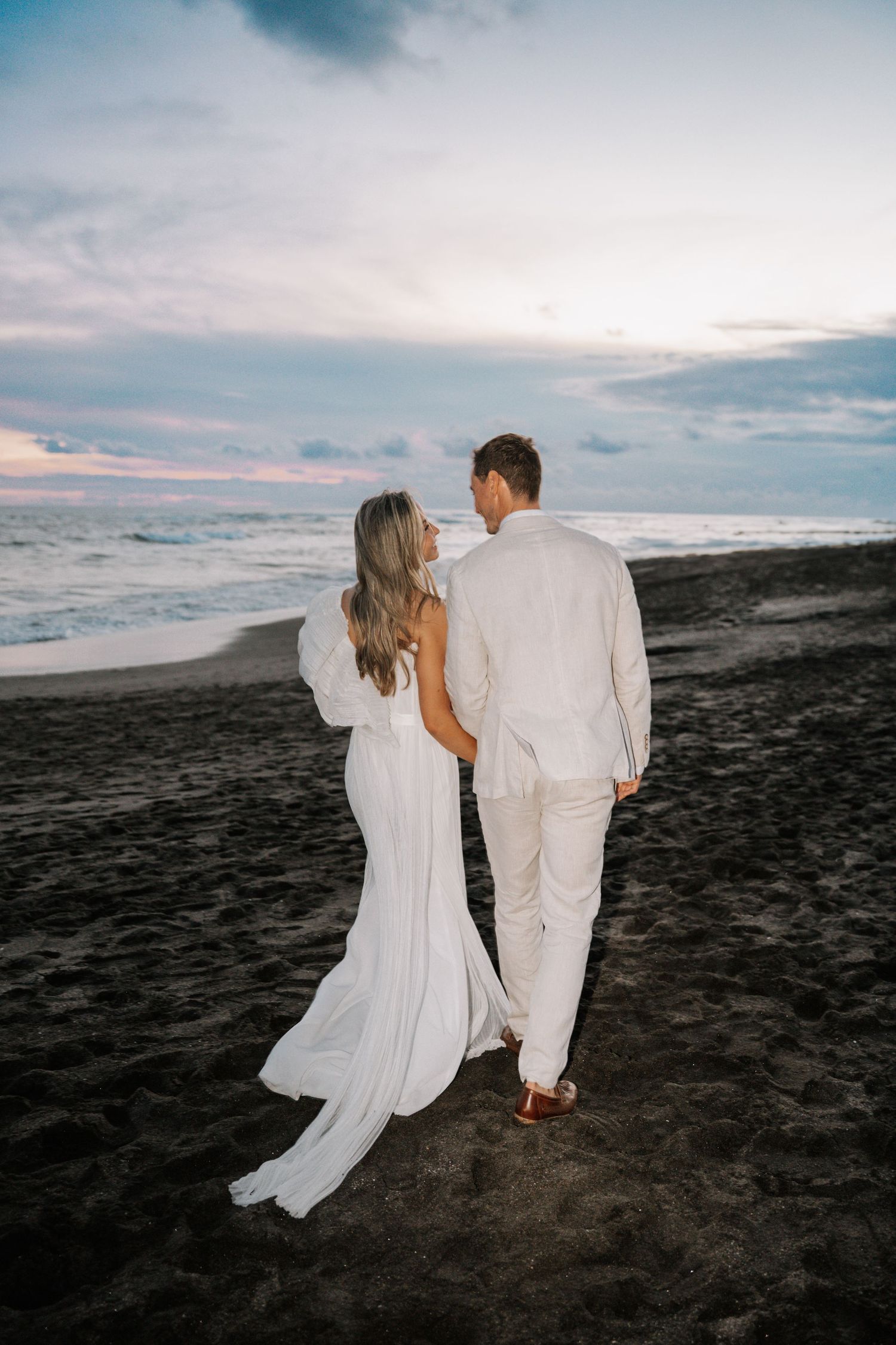 A couple in white attire walking together on a black sand beach at sunset with ocean waves in the background.