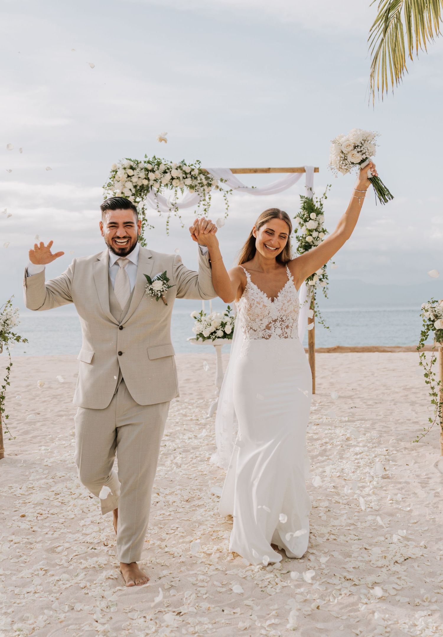 A joyful wedding celebration with a couple jumping together on a tropical beach with palm trees and ocean backdrop.