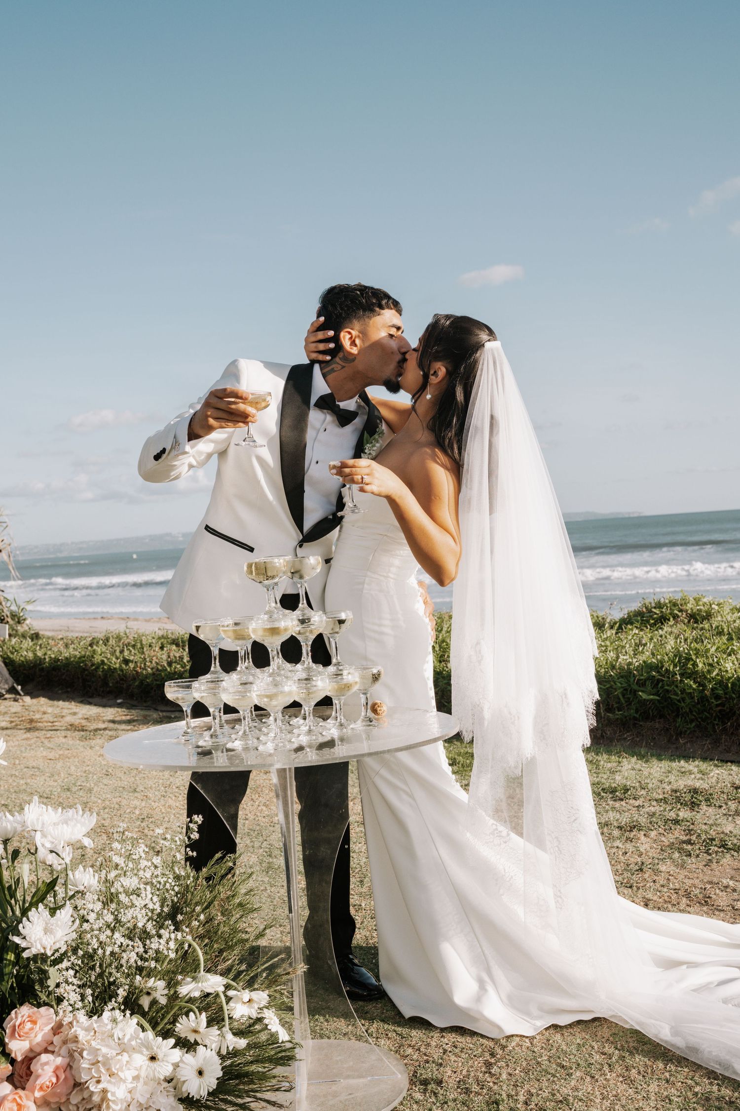 A newlywed couple shares a romantic moment by a champagne tower at their oceanside wedding celebration.