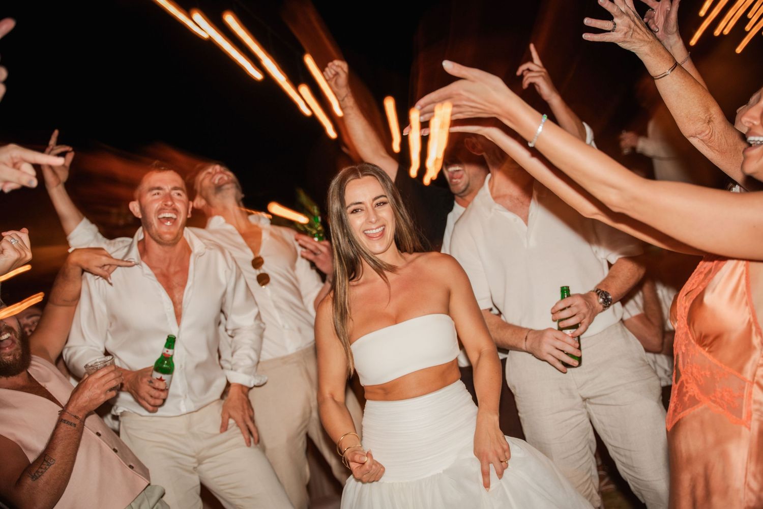 Wedding party celebration at night with guests dancing and raising their hands under light flares.