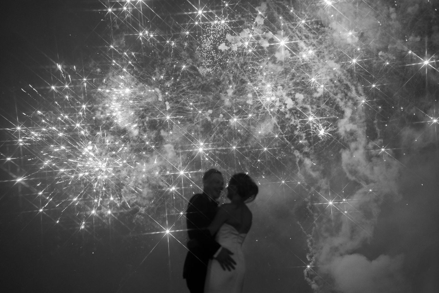Silhouetted couple embraces while watching fireworks light up the night sky in black and white.