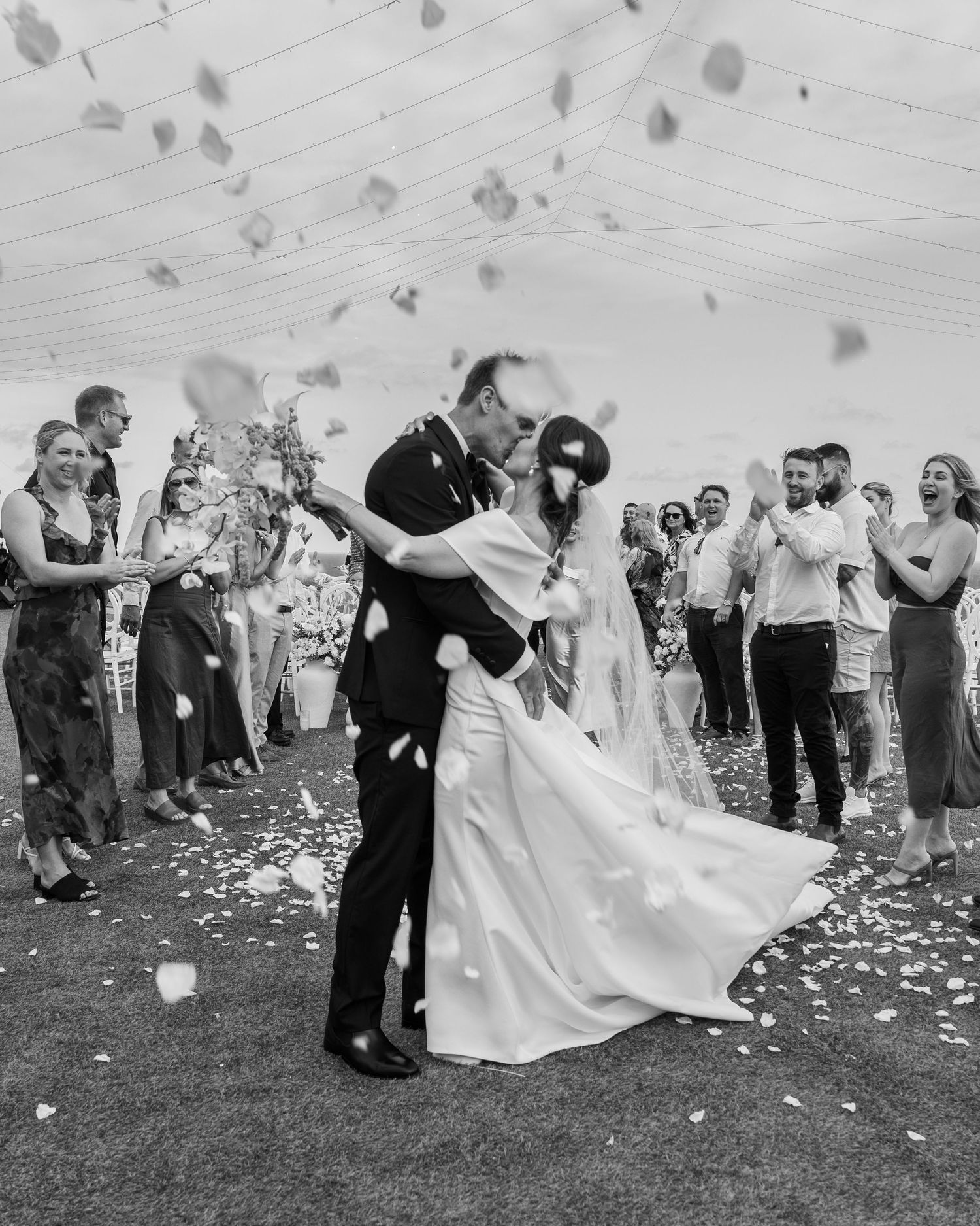 Wedding couple shares a romantic dip kiss while guests shower them with flower petals during an outdoor celebration.