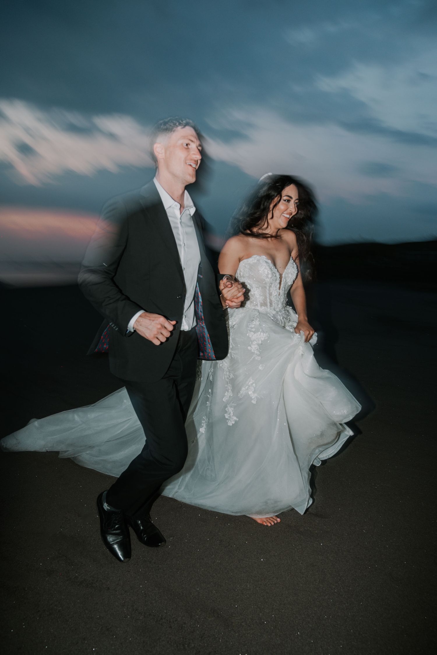 A wedding couple dances and poses together on a dark beach at dusk with dramatic cloudy skies in the background.