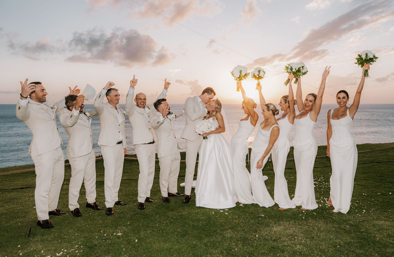 Wedding party in white attire celebrating outdoors by the ocean at sunset, bride and groom kissing while guests cheer.