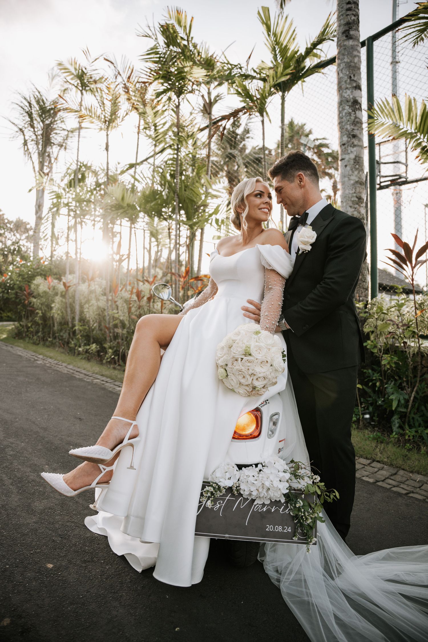 Romantic wedding photo with couple posed against scooter and palm trees.