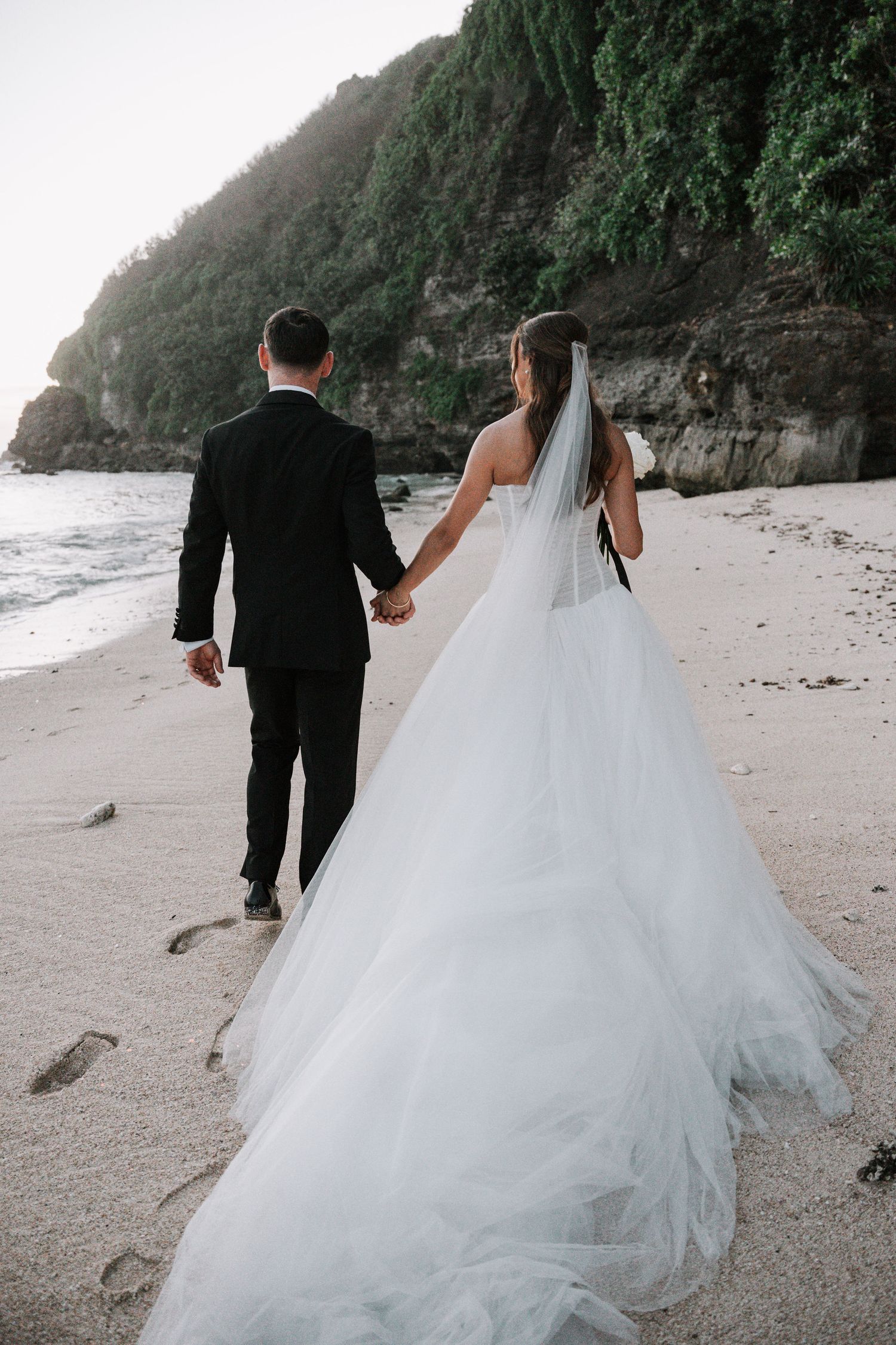 A couple in wedding attire walks along a scenic rocky beach with dramatic cliffs in the background.