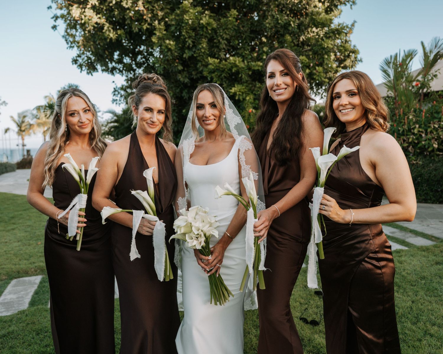 Bride in white gown with four bridesmaids in brown satin dresses holding white floral bouquets outdoors.