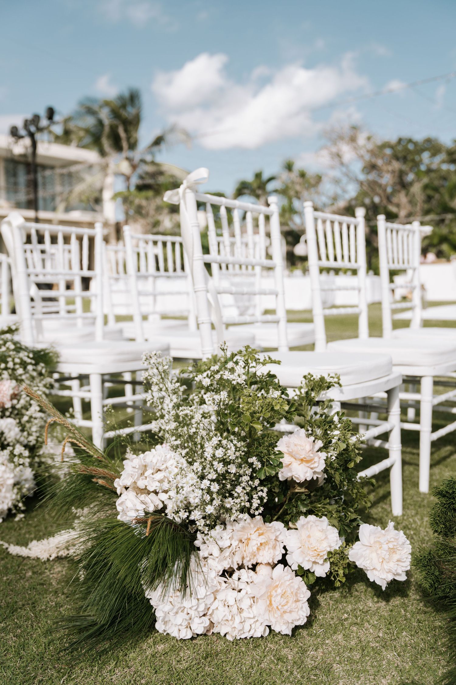 White wooden chairs arranged outdoors with delicate floral arrangements lining the aisle for an elegant garden wedding ceremony.