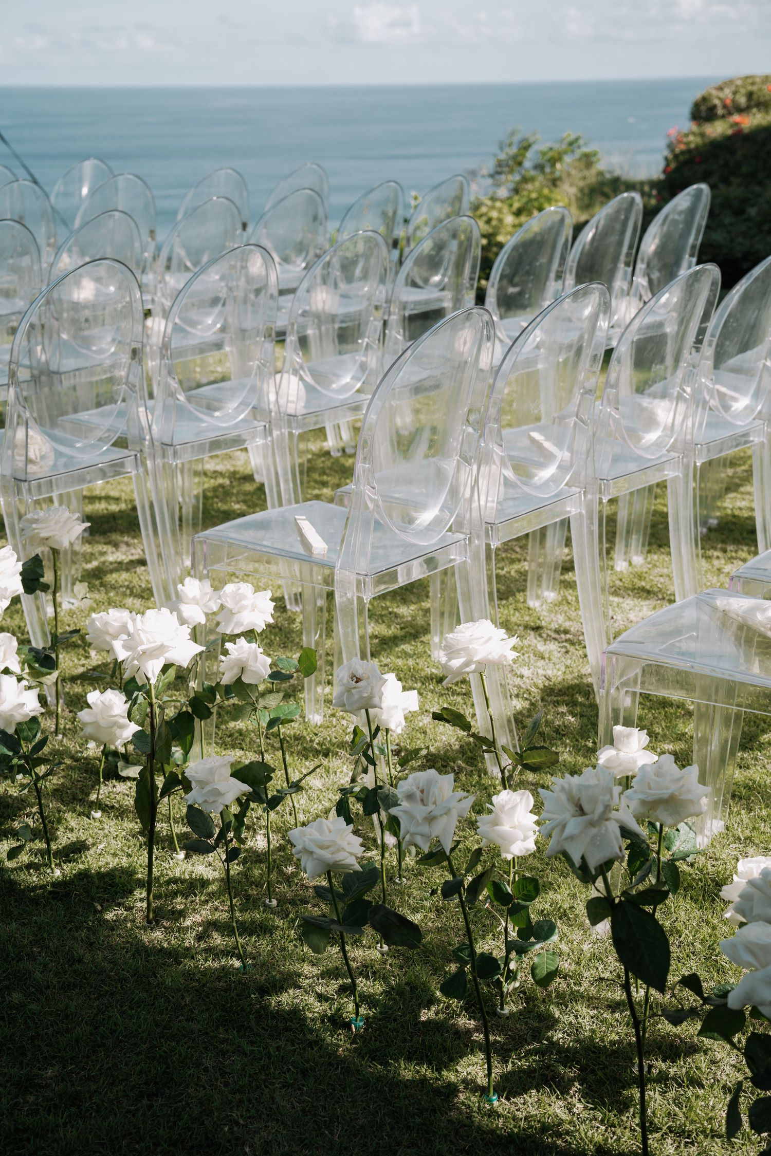 Clear acrylic chairs line up along a scenic oceanfront wedding ceremony setup with white floral arrangements.