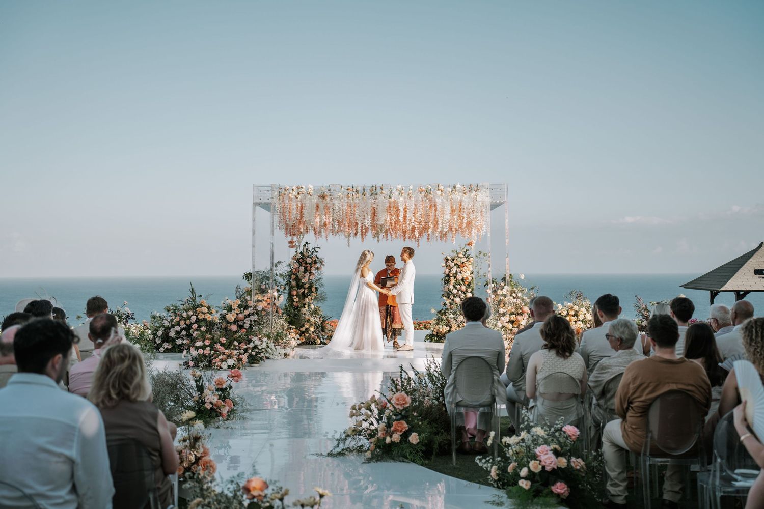 Oceanfront wedding ceremony setup with floral arch and white chairs against a blue sky.