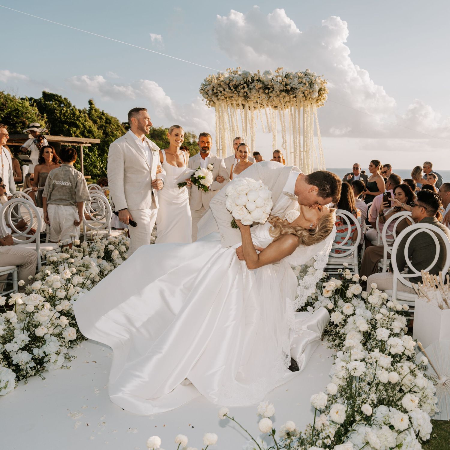 Groom dips bride in white gown at outdoor wedding ceremony with floral arch and white roses.