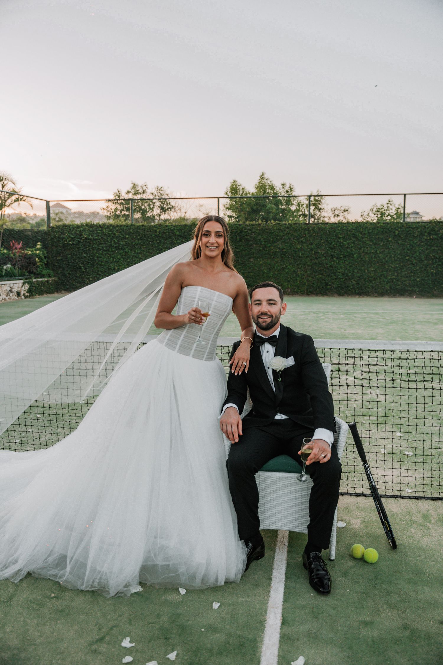 A bride and groom pose playfully on a tennis court with tennis balls and flowing white veil during their wedding photoshoot.