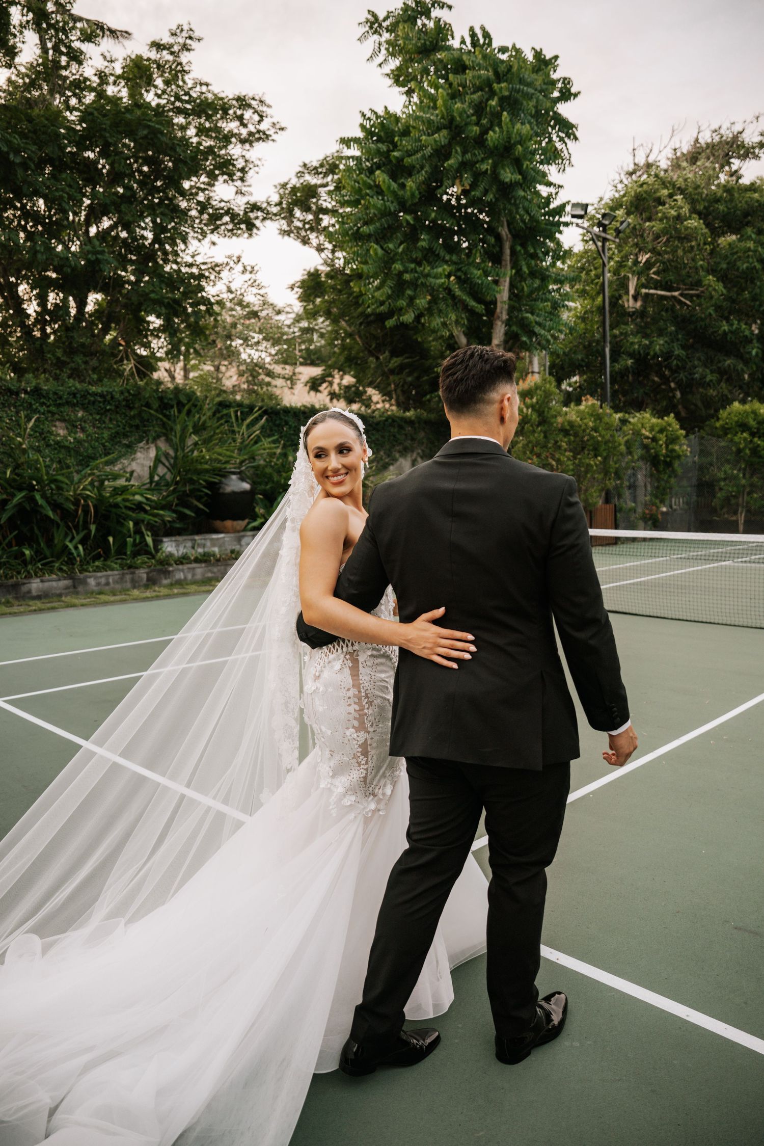 Newlywed couple shares a playful moment on a green tennis court with bride's veil flowing in the breeze.