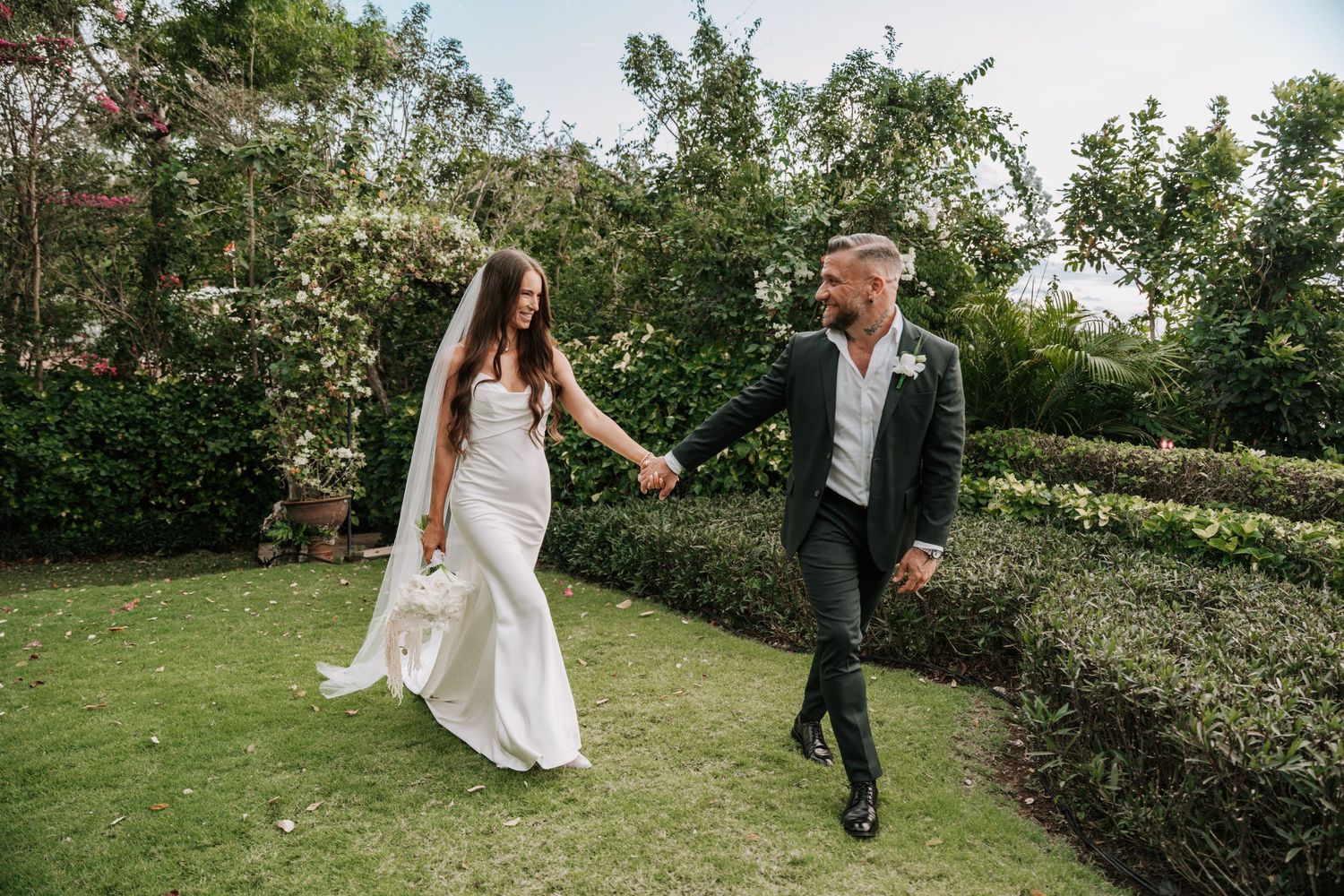 Wedding couple walks hand in hand through a lush green garden during outdoor summer celebration.