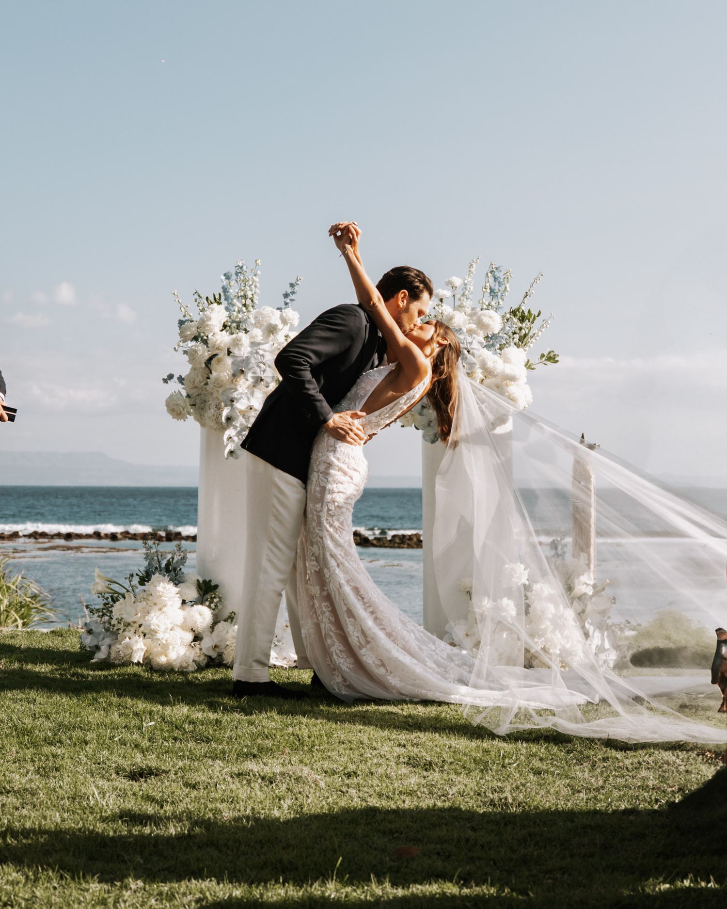 A couple in wedding attire dips and poses dramatically on a grassy hilltop overlooking the ocean with flowing white veil.