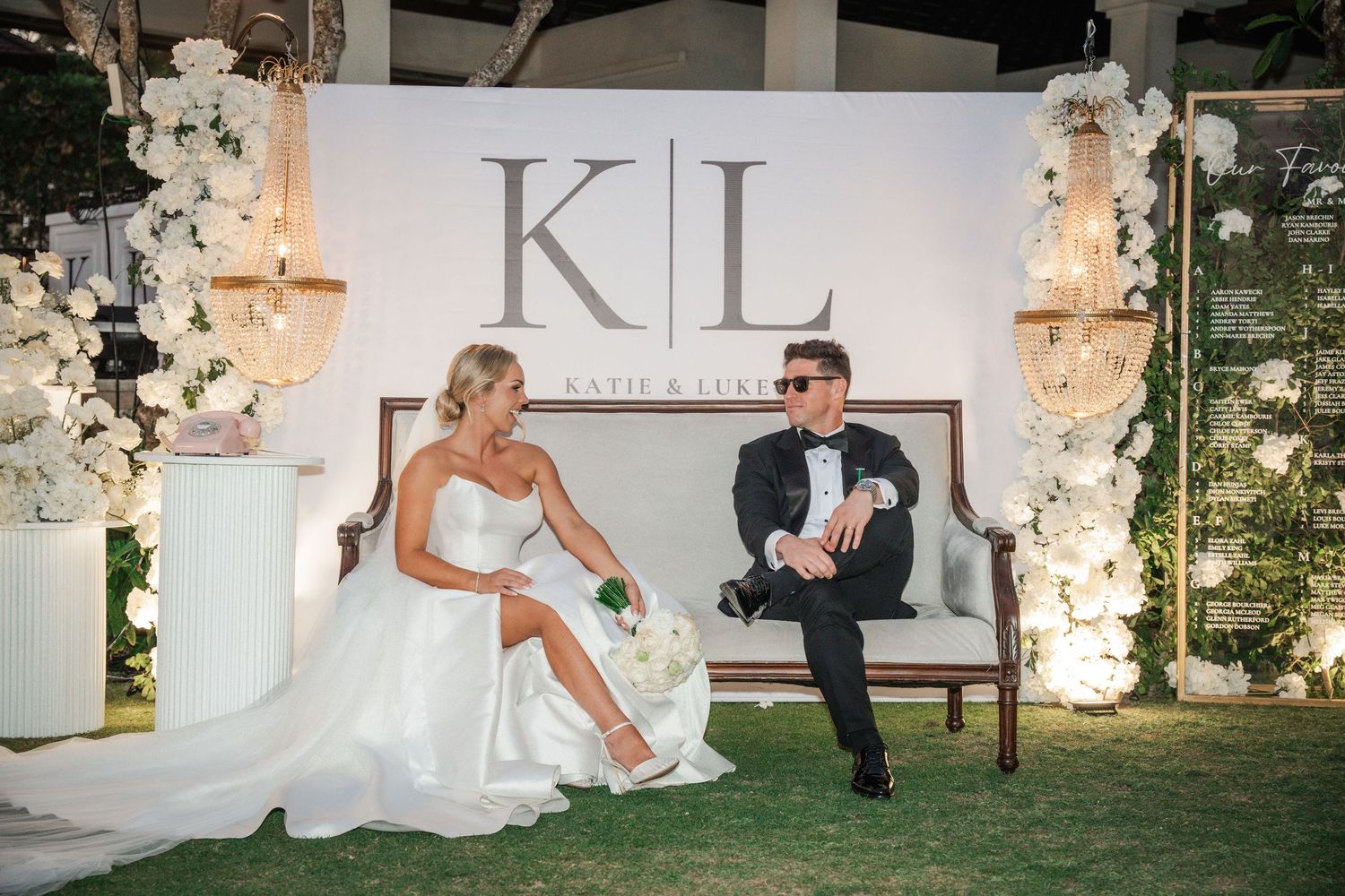 Wedding couple relaxing on a bench under elegant 'KL' monogram display with floral decor.