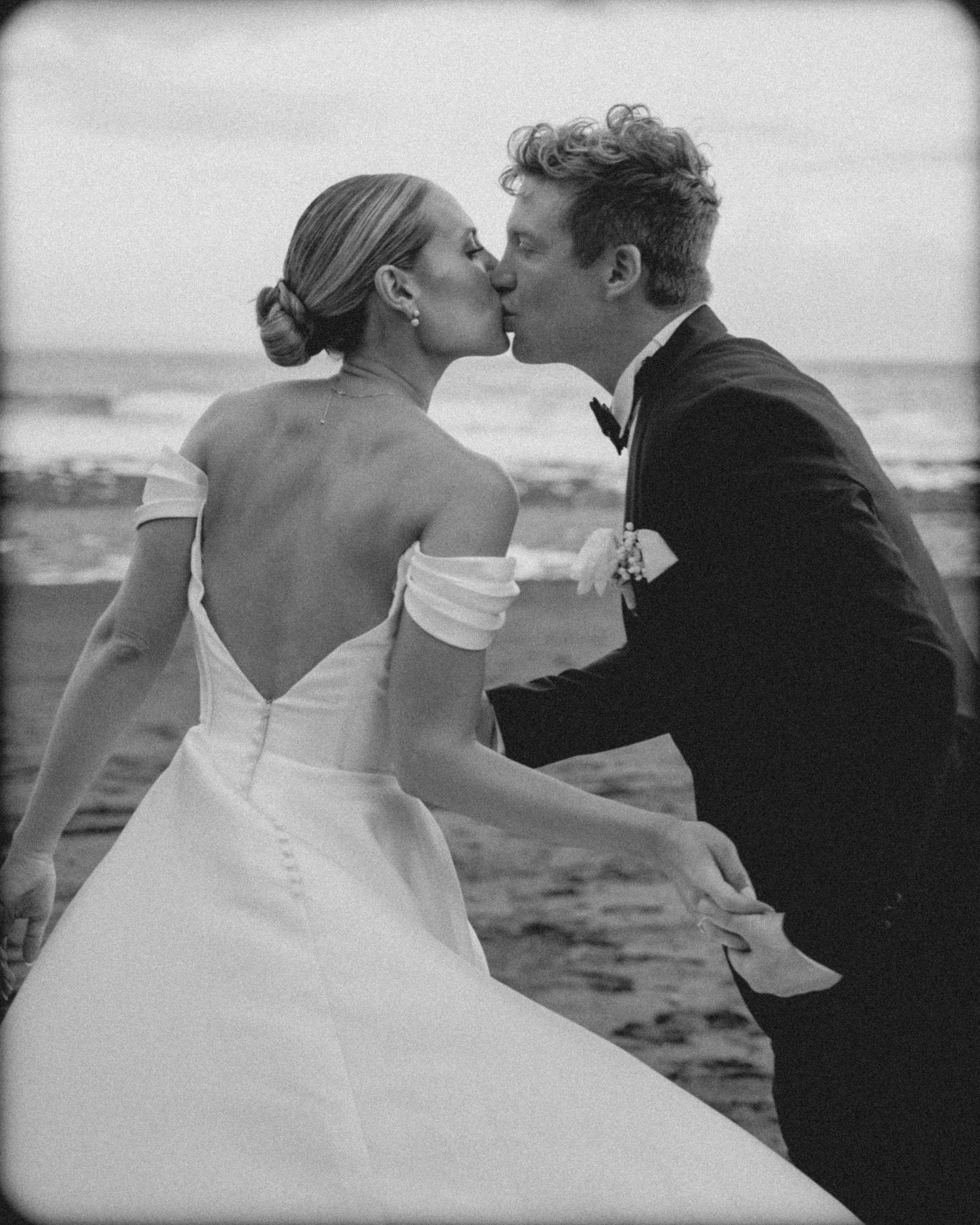 A romantic black and white beach wedding photo shows a couple sharing a kiss in formal attire by the ocean.