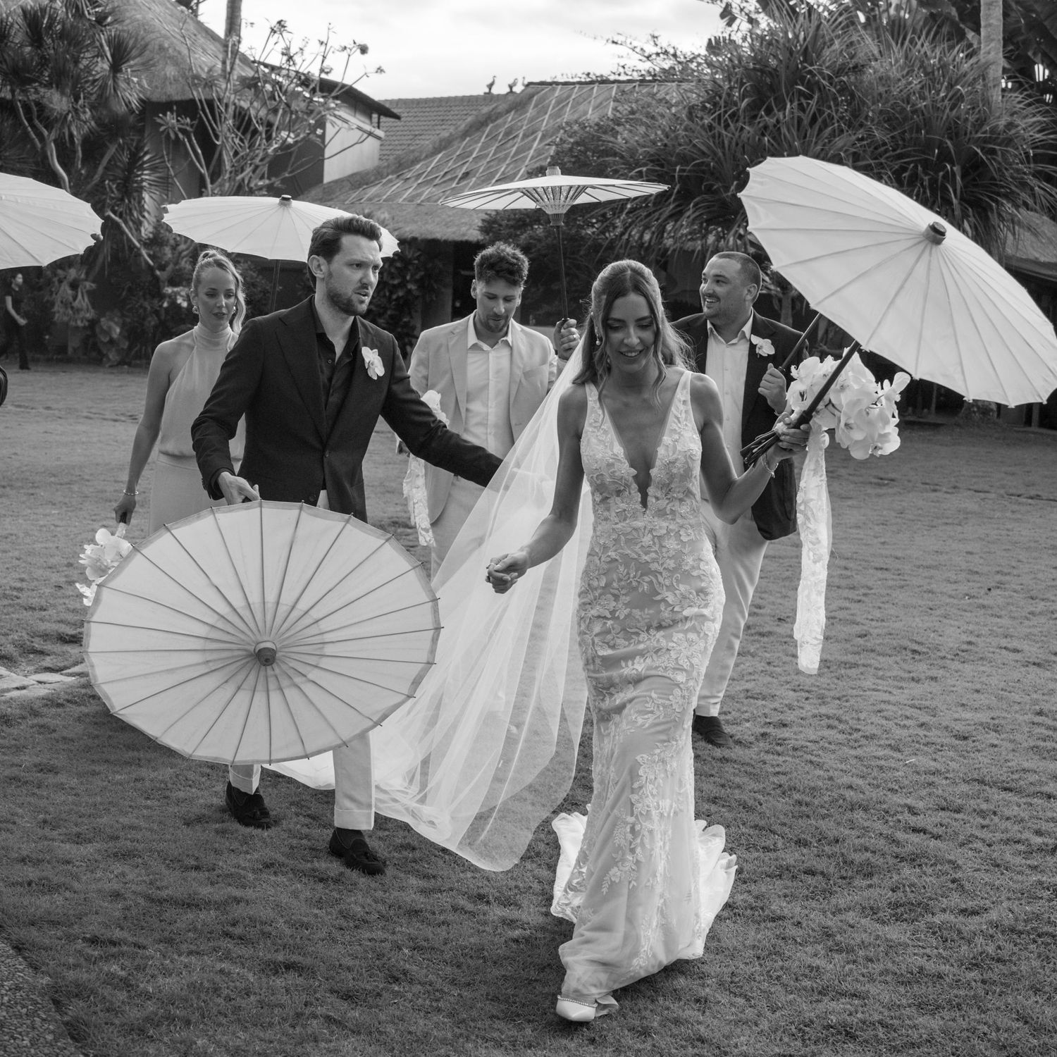 Wedding party walks across grass holding white paper parasols during an outdoor celebration.