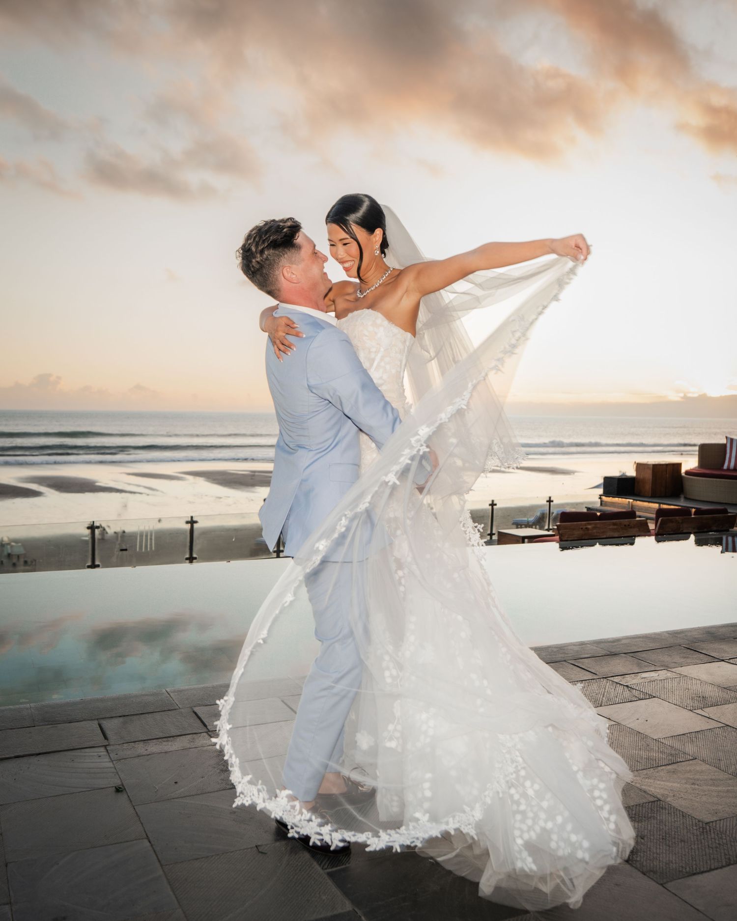 Newlywed couple shares romantic sunset moment on a beachside terrace as her veil and dress flow in the ocean breeze.