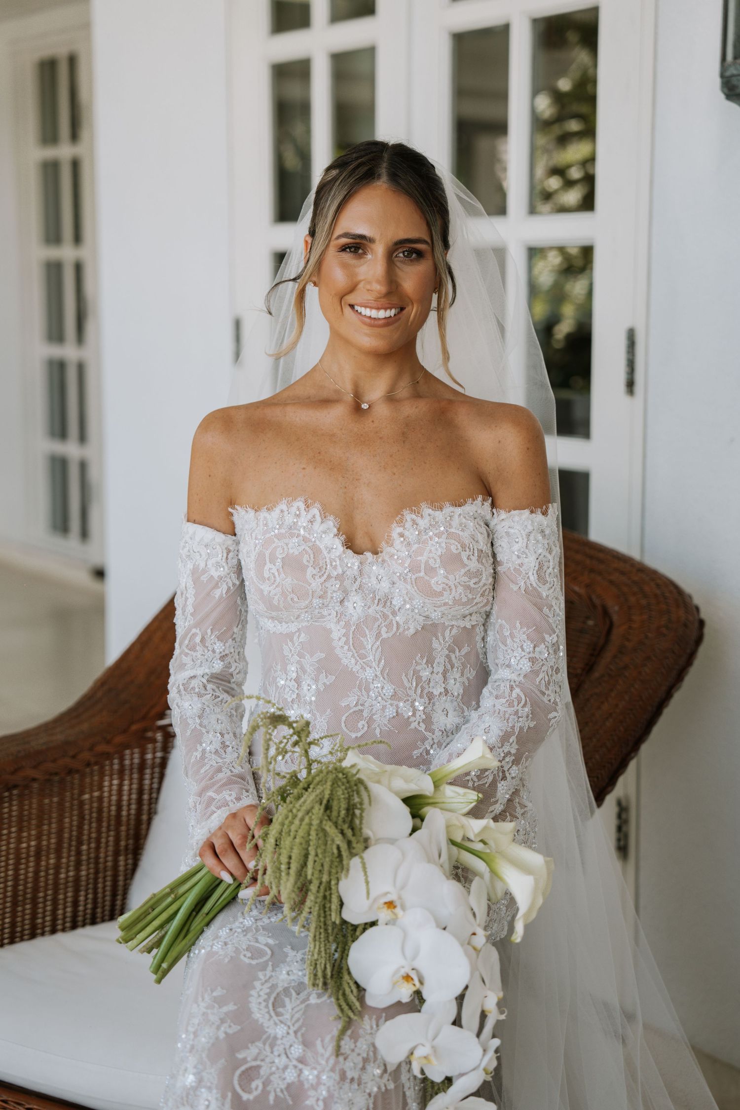 Bride in off-shoulder lace wedding dress holds elegant white orchid bouquet on porch with wicker furniture.