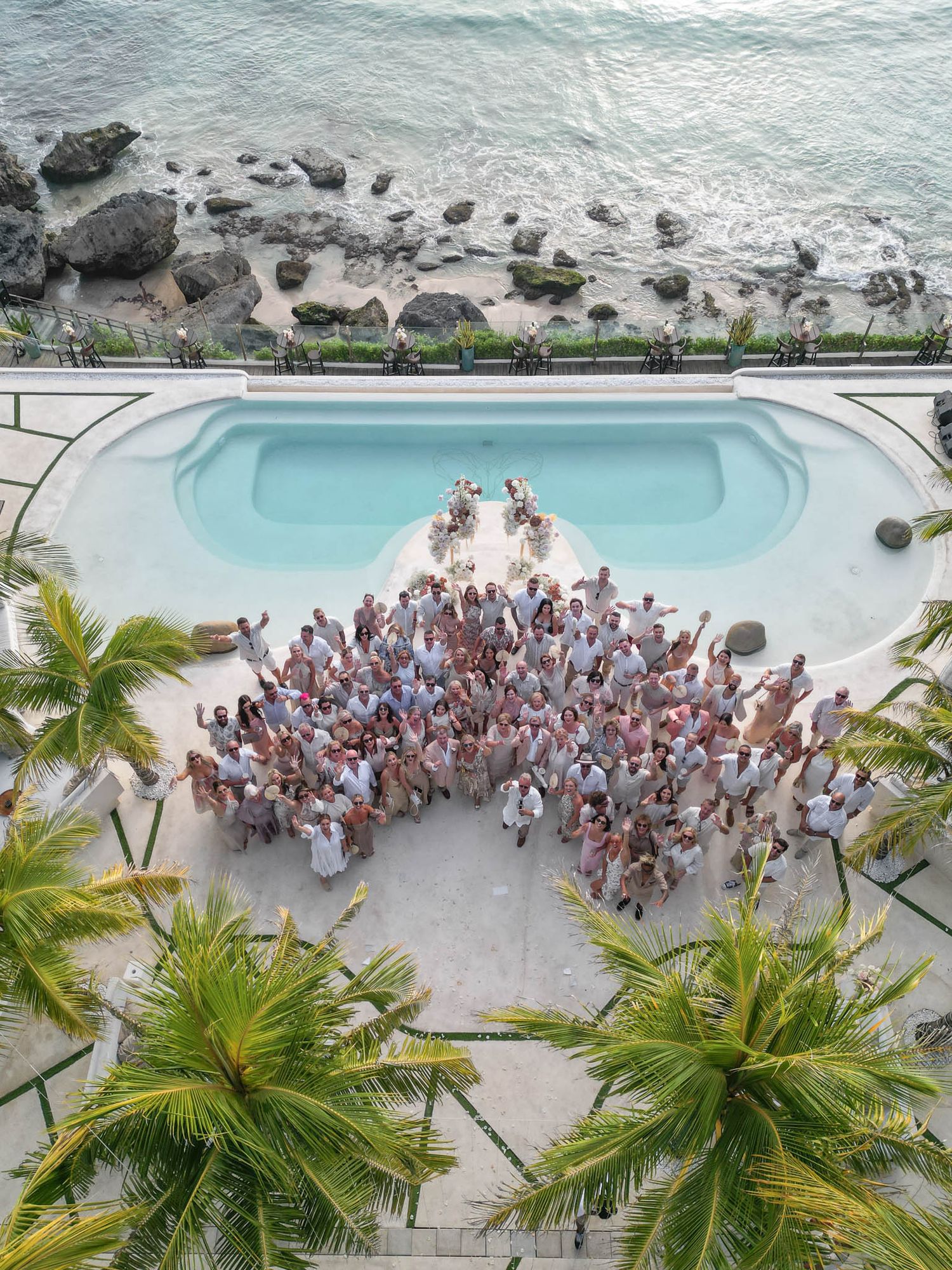 Large group of wedding guests gathered around a tropical resort pool with palm trees and ocean views.