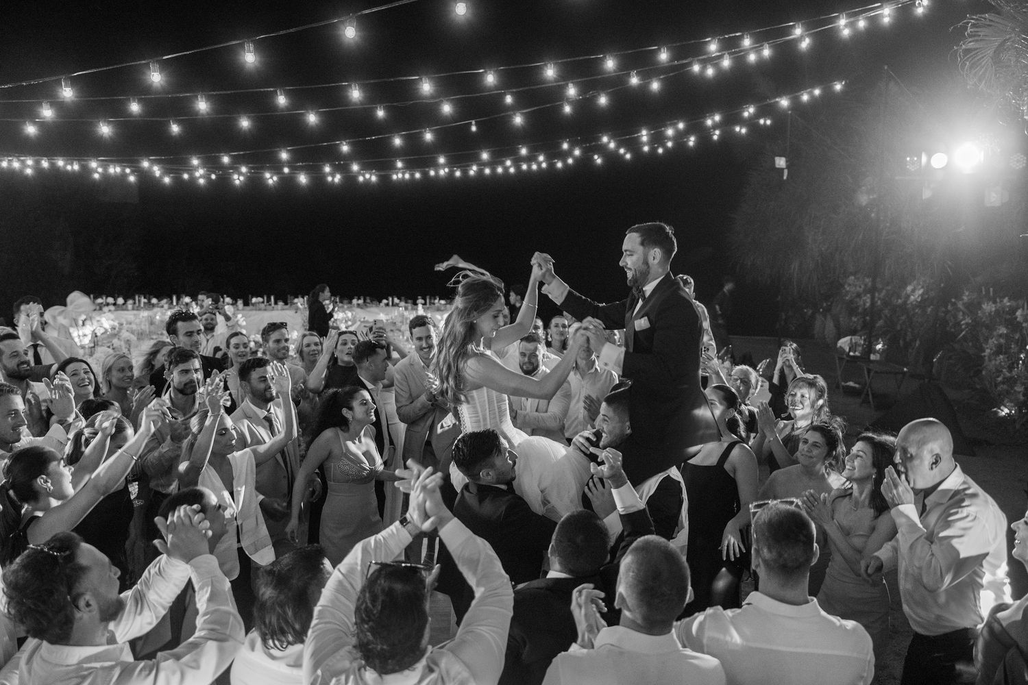 Wedding reception crowd dances under twinkling string lights at an outdoor evening celebration.