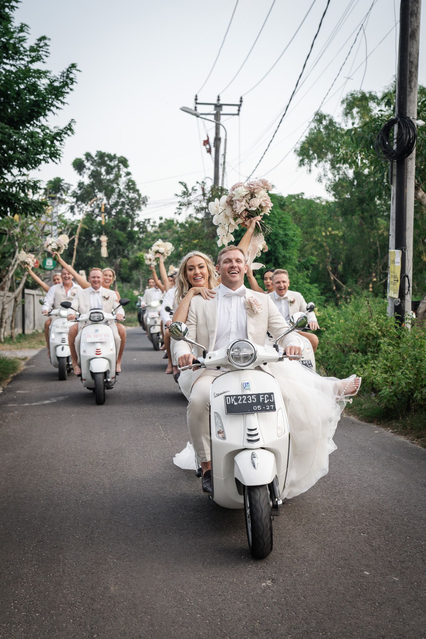 Wedding party rides white Vespa scooters down a tree-lined street in celebratory procession.