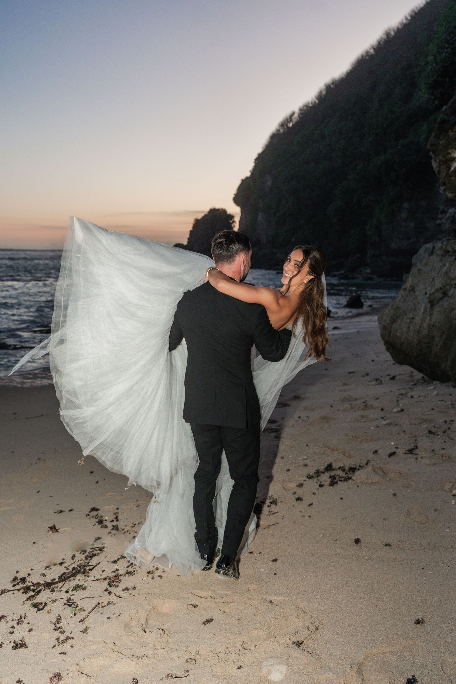 A sunset beach wedding photo with waves crashing against rocky cliffs in the background.