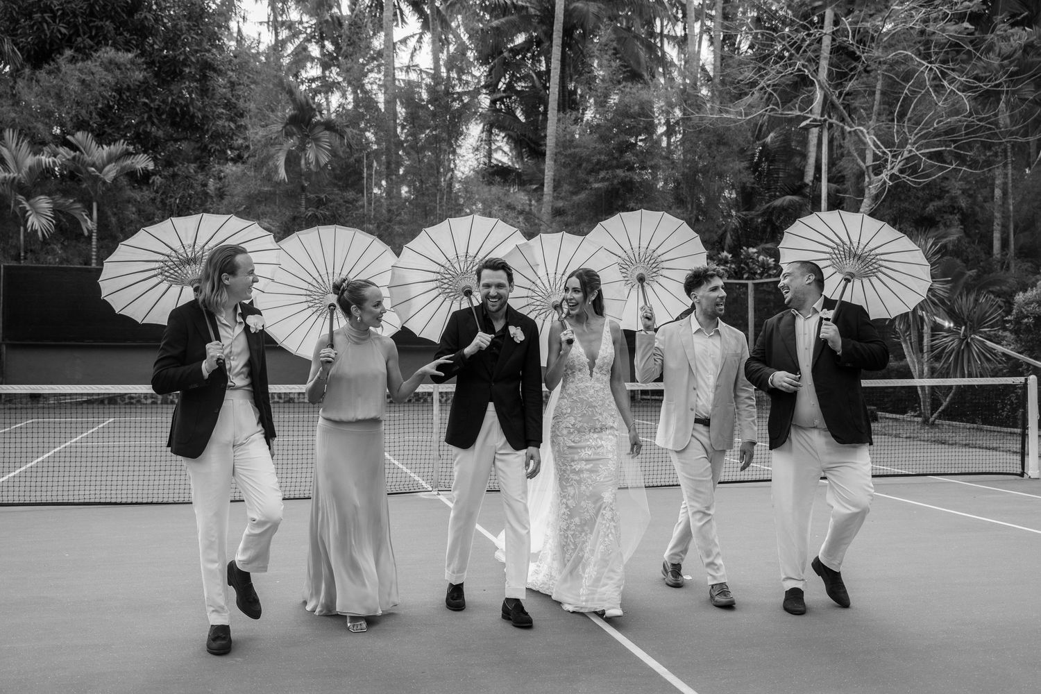 Wedding party members in white attire pose with paper parasols on a tennis court surrounded by palm trees.