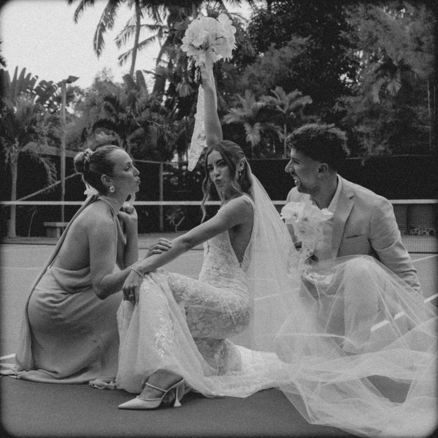 A candid black and white wedding moment captured on an outdoor tennis court with tropical scenery.