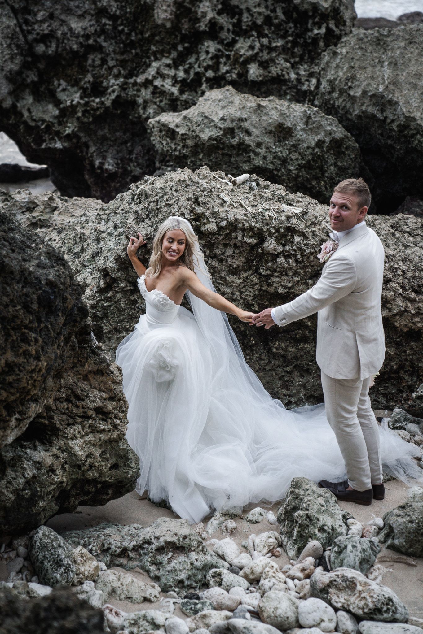 Wedding couple in white attire playfully posing among dramatic rocky coastal formations.