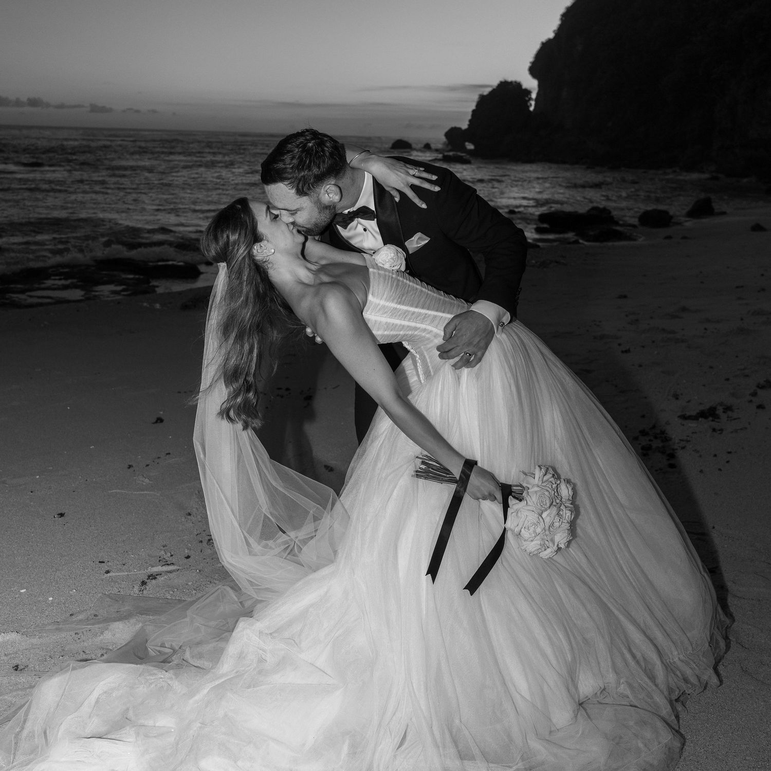 A series of black and white beach wedding photos showing a bride in a flowing white gown posing on the sand at sunset.