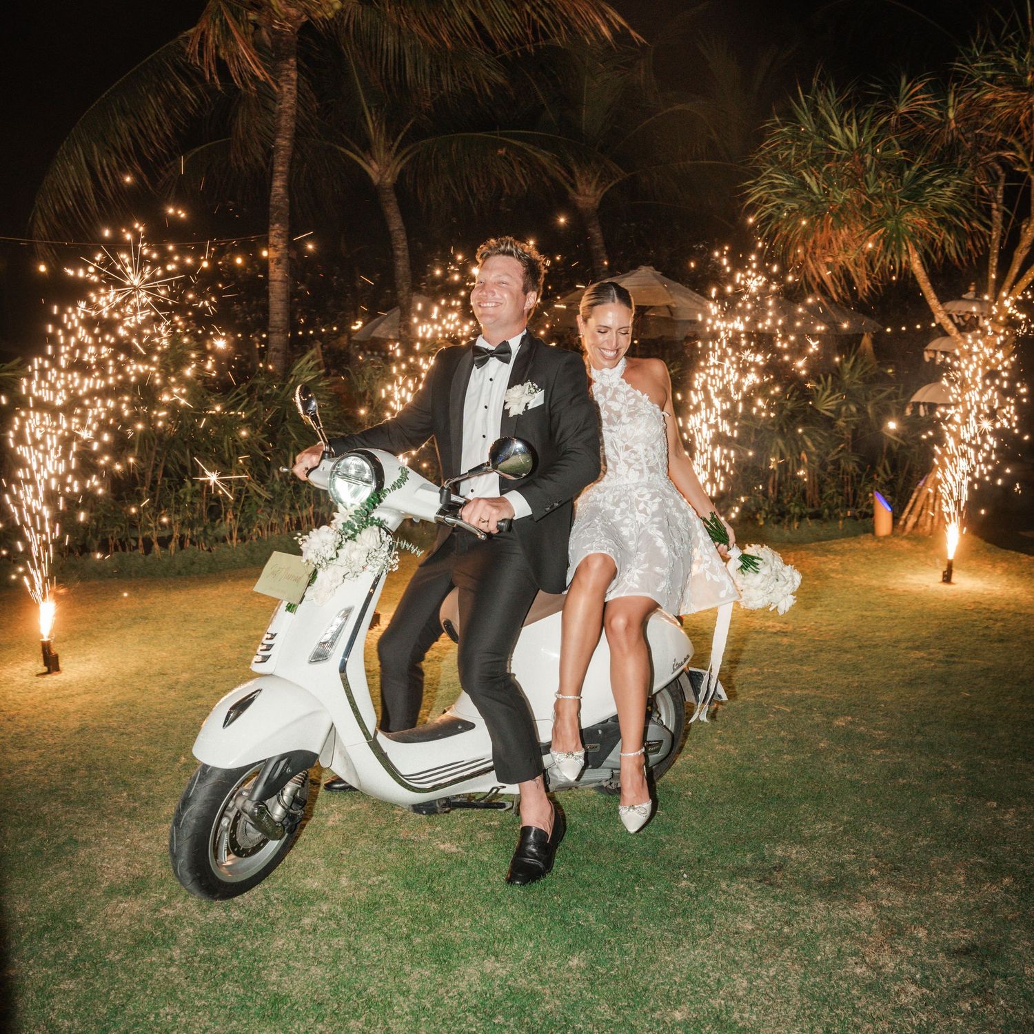 Couple sitting on a vintage white Vespa scooter at night with sparklers and twinkle lights creating a romantic atmosphere.