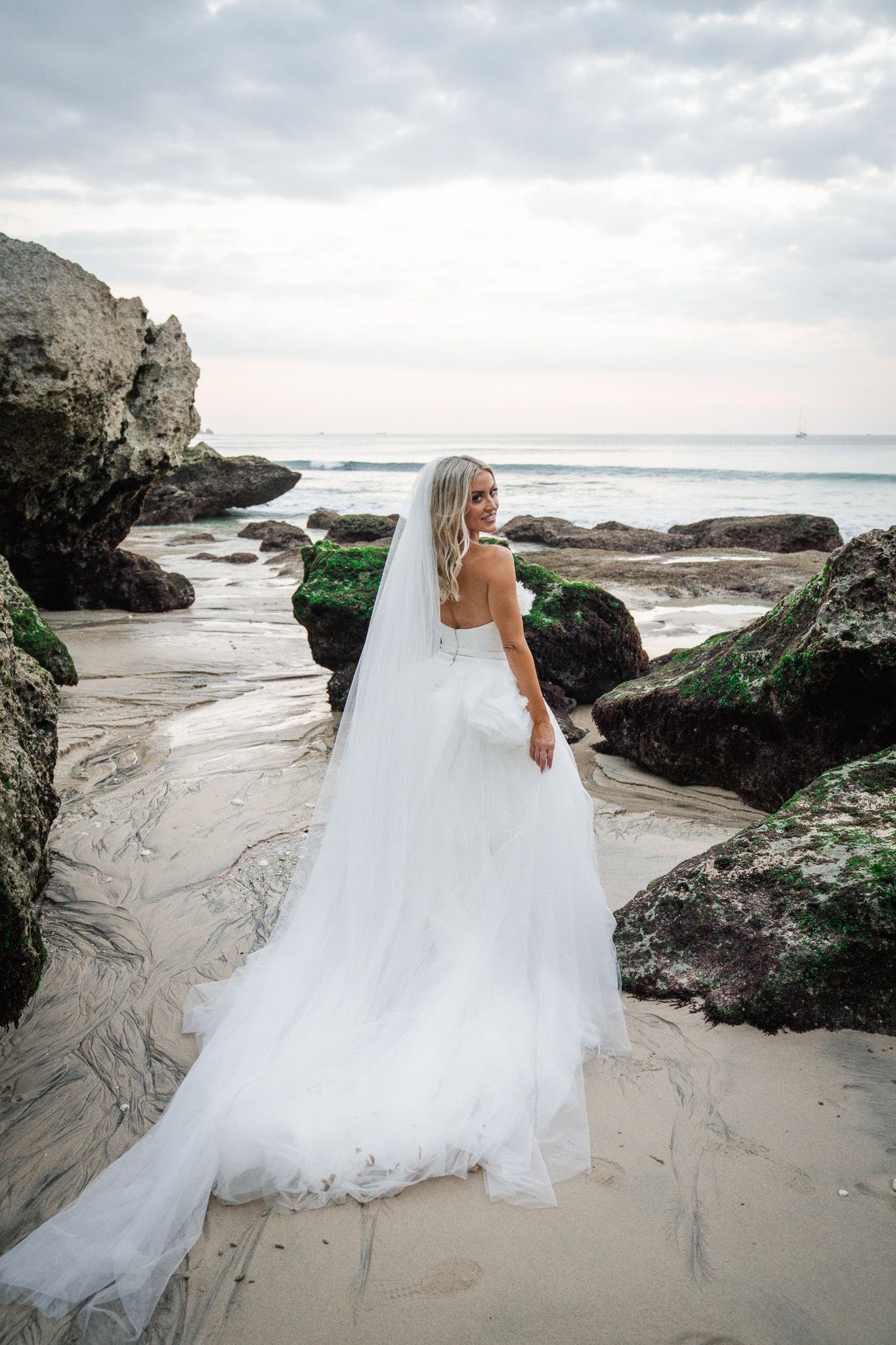 A bride in a flowing white gown poses on a rocky beach with ocean waves and cliffs in the background.