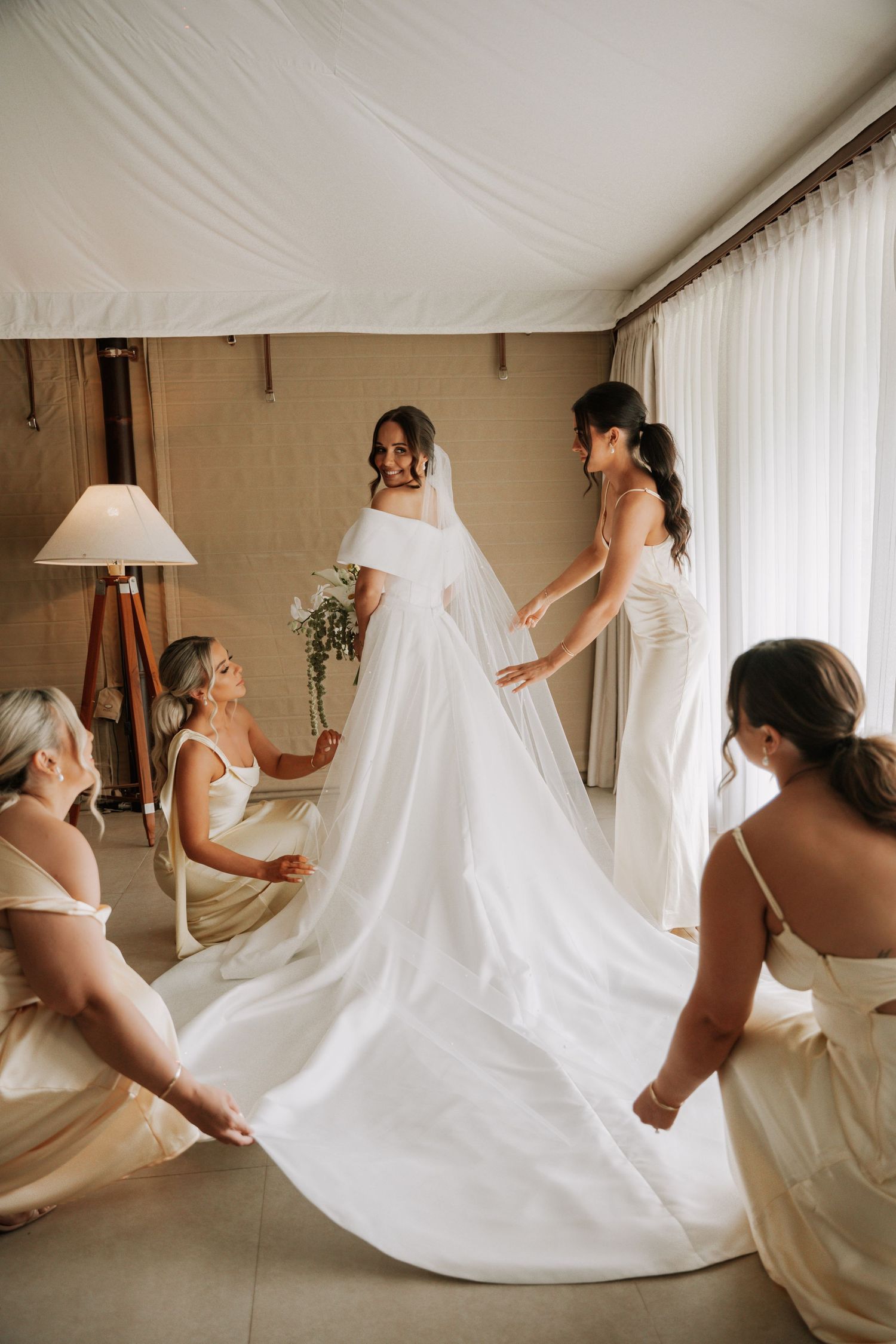 Bridesmaids in cream dresses help arrange the long train of an elegant white wedding gown in a sunlit room.