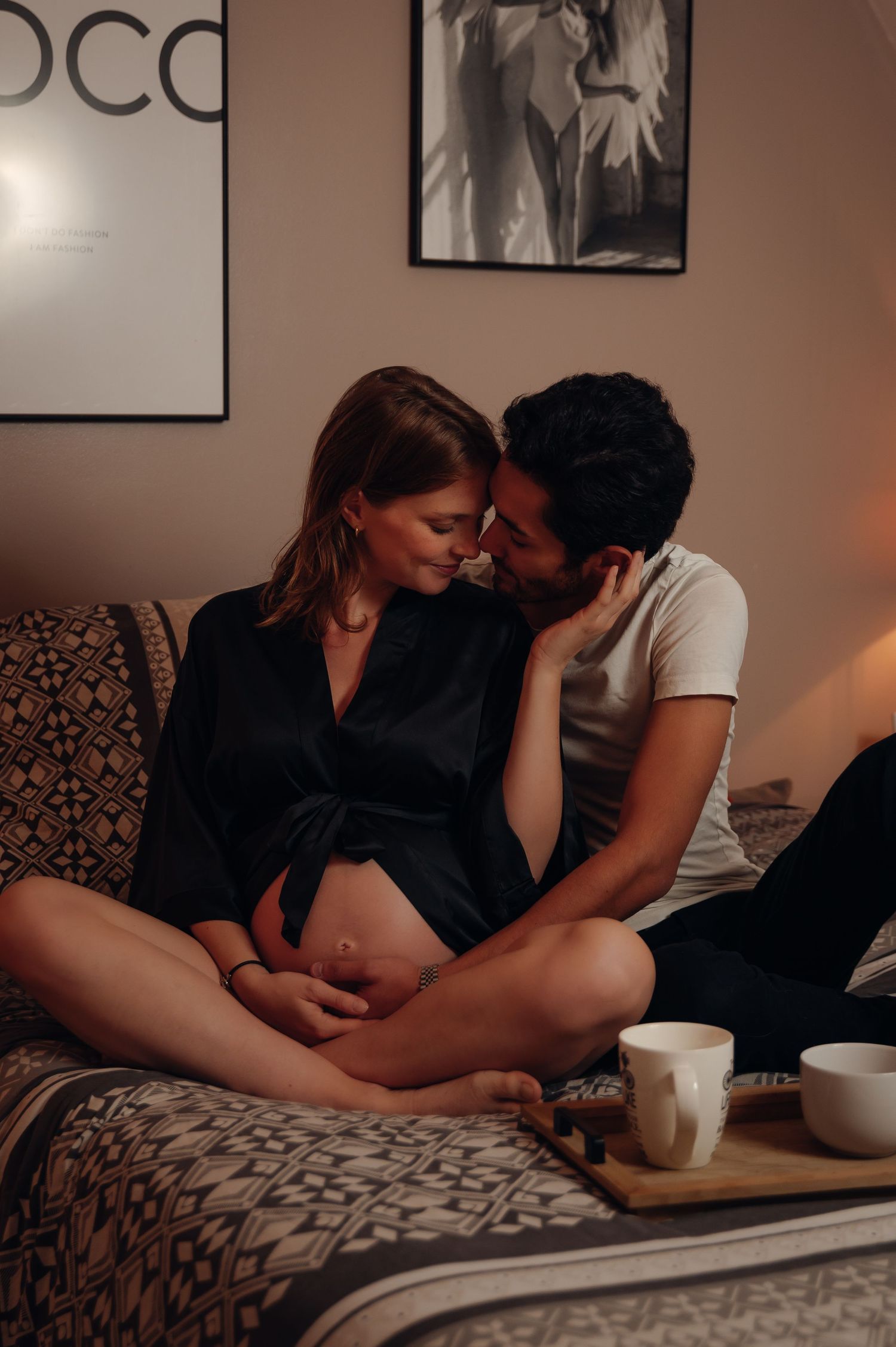 Pregnant couple sharing an intimate moment on a cozy bed, woman in black silk robe cradling her baby bump.
