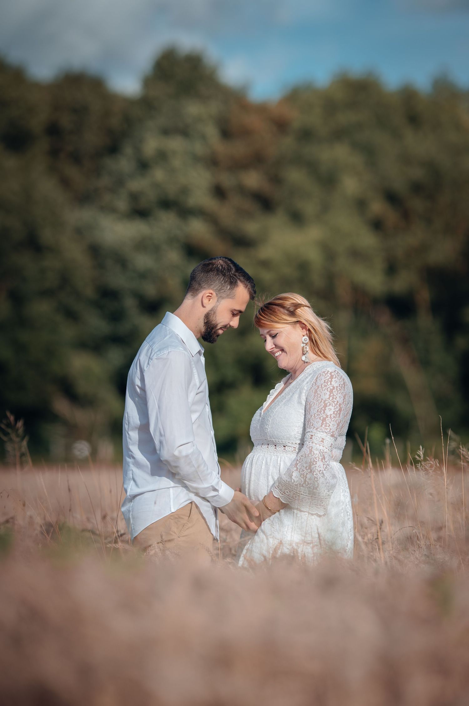 Pregnant couple holding hands in a field, smiling down at baby bump, wearing white outfits with green trees behind.