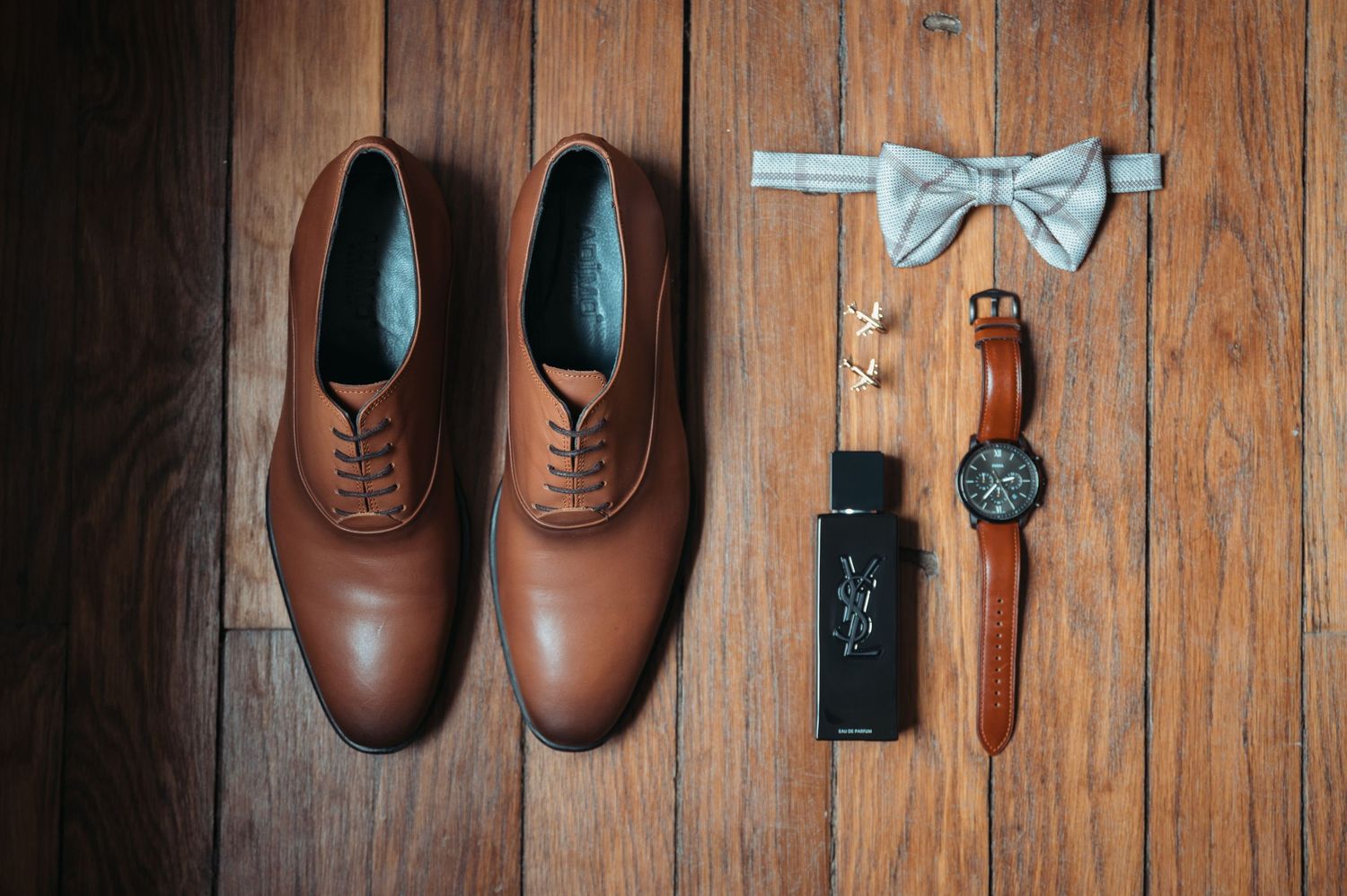 Flat lay of brown leather oxford shoes, bow tie, watch, and cologne bottle on wooden floor.