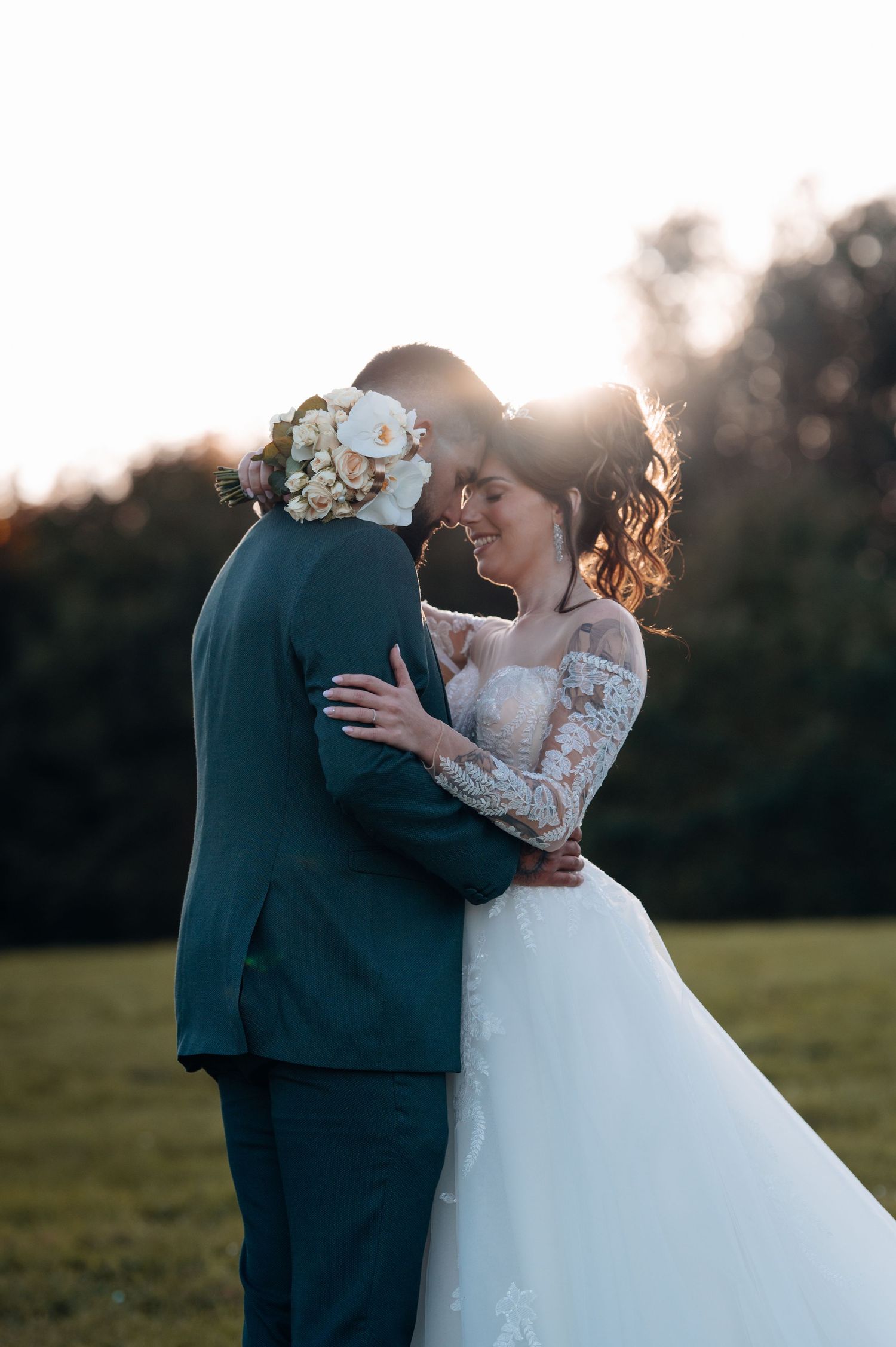 Happy bride in lace gown embracing groom with floral bouquet at golden hour sunset in green field.