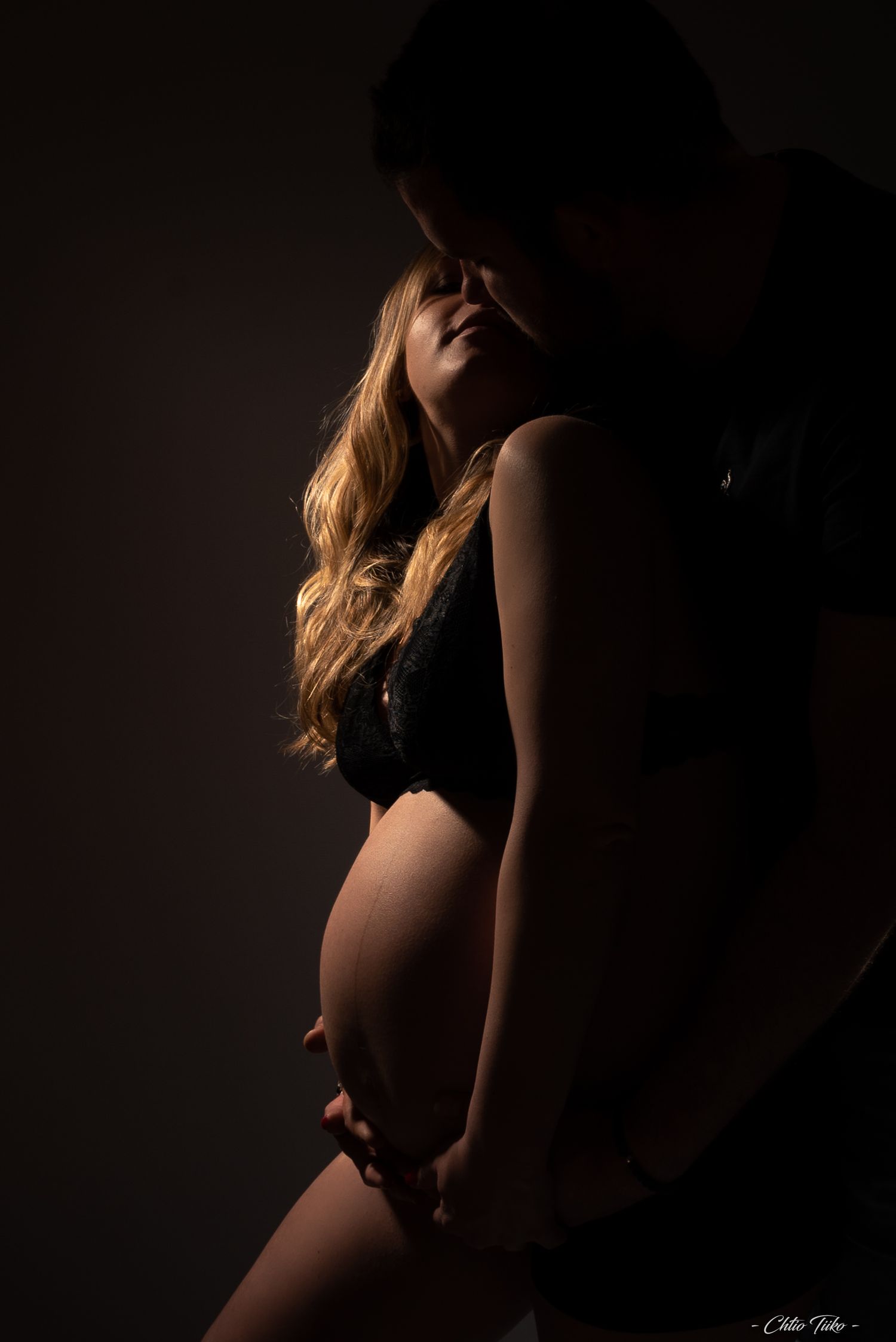 Silhouette of pregnant woman in black lingerie with partner in dramatic low-key maternity portrait.