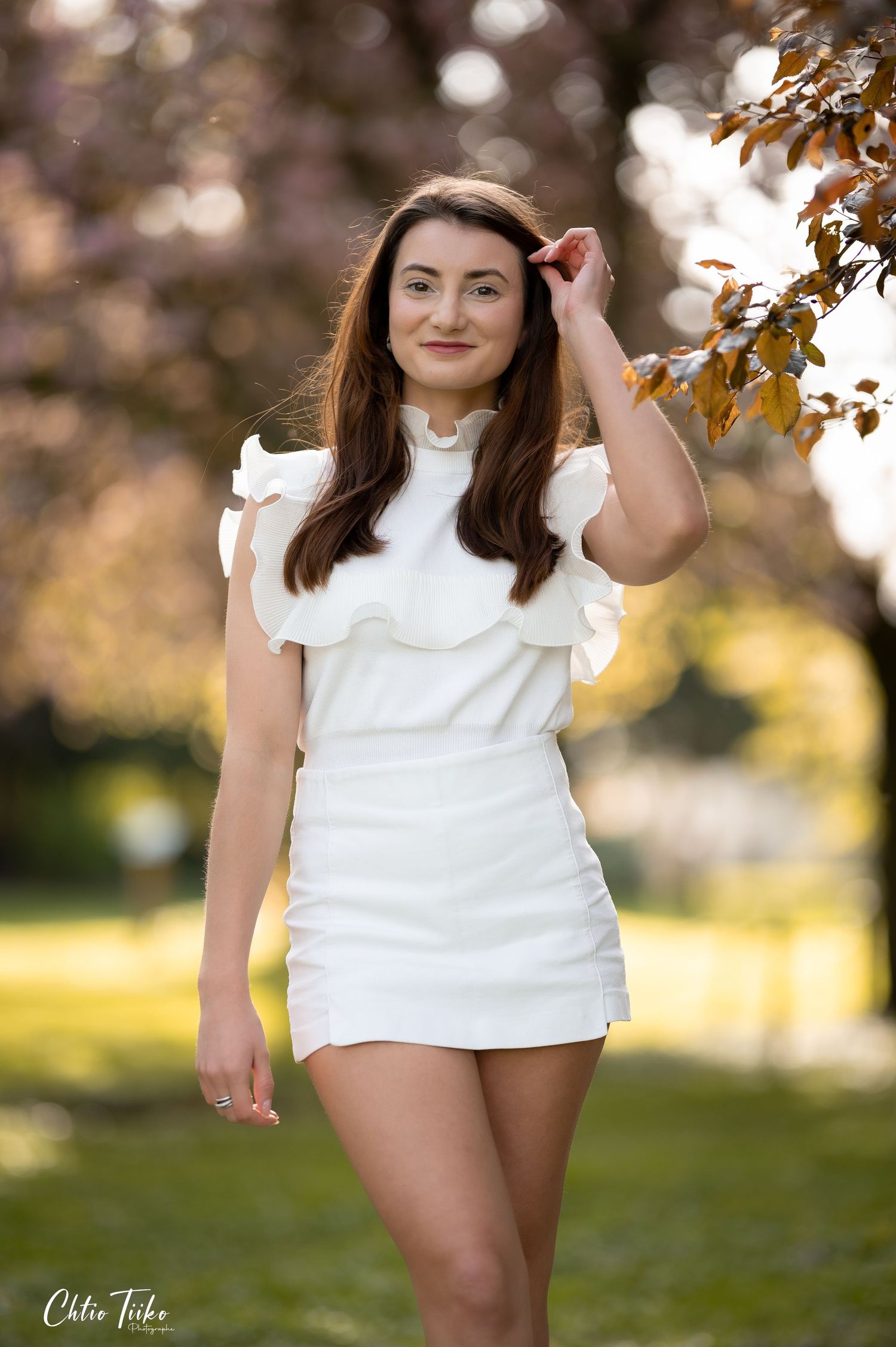 Young woman in white ruffled mini dress poses outdoors near cherry blossom tree in a sunny park.