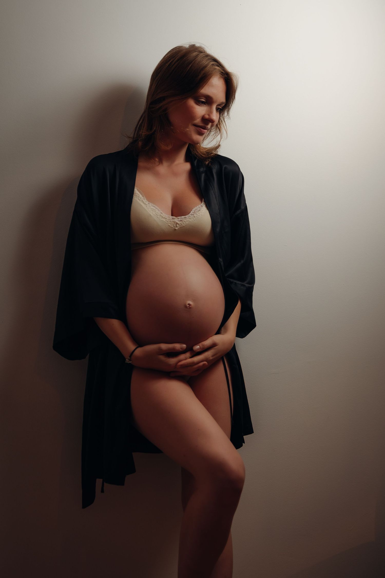 Pregnant woman in beige lingerie and black robe cradling her baby bump against a neutral wall.